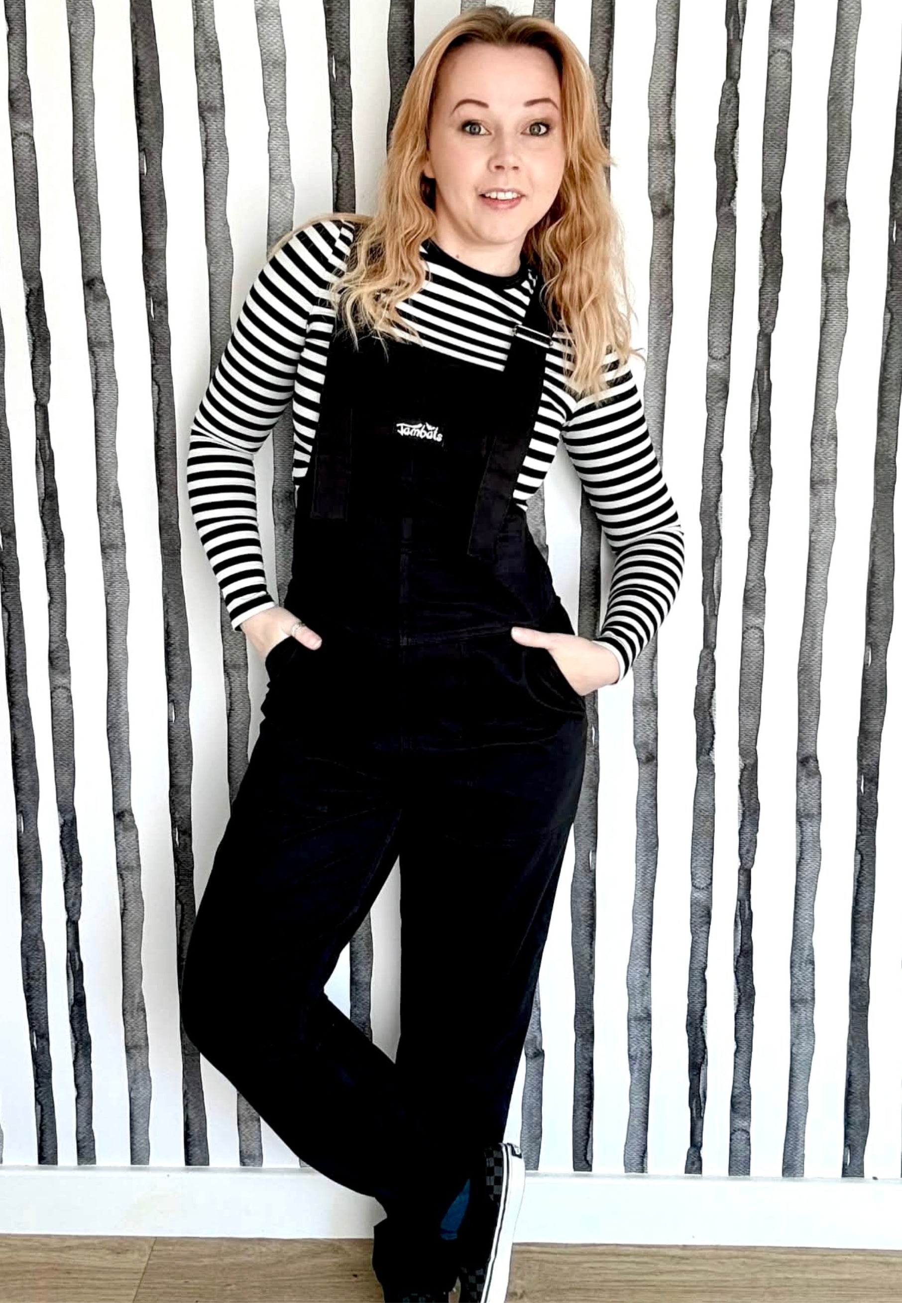 Woman wearing black dungarees overalls and a striped shirt against a quirky striped wall.