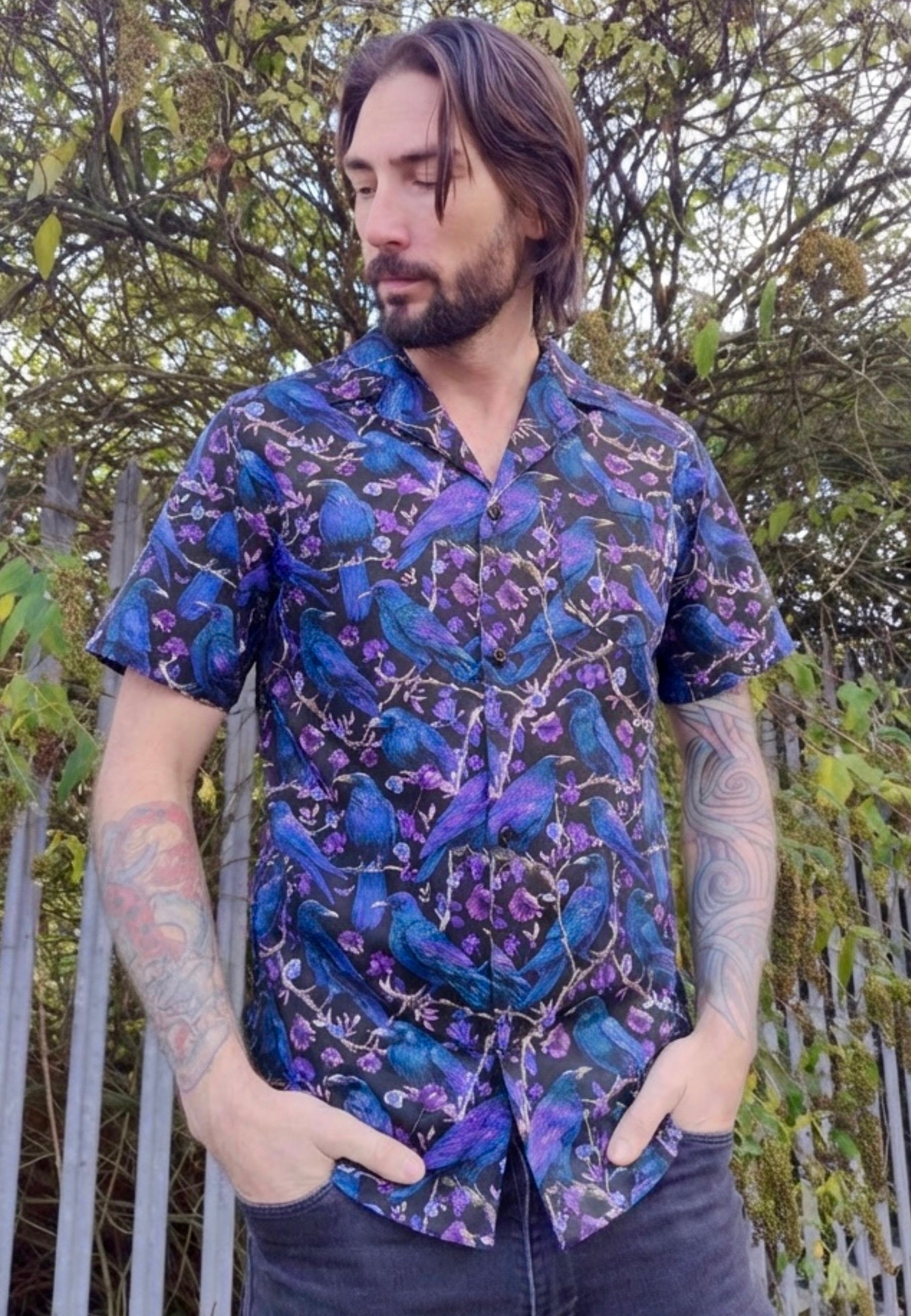 Man wearing a shirt with a print of deep purple and blue ravens and crows on a black background. Photo is by some trees 