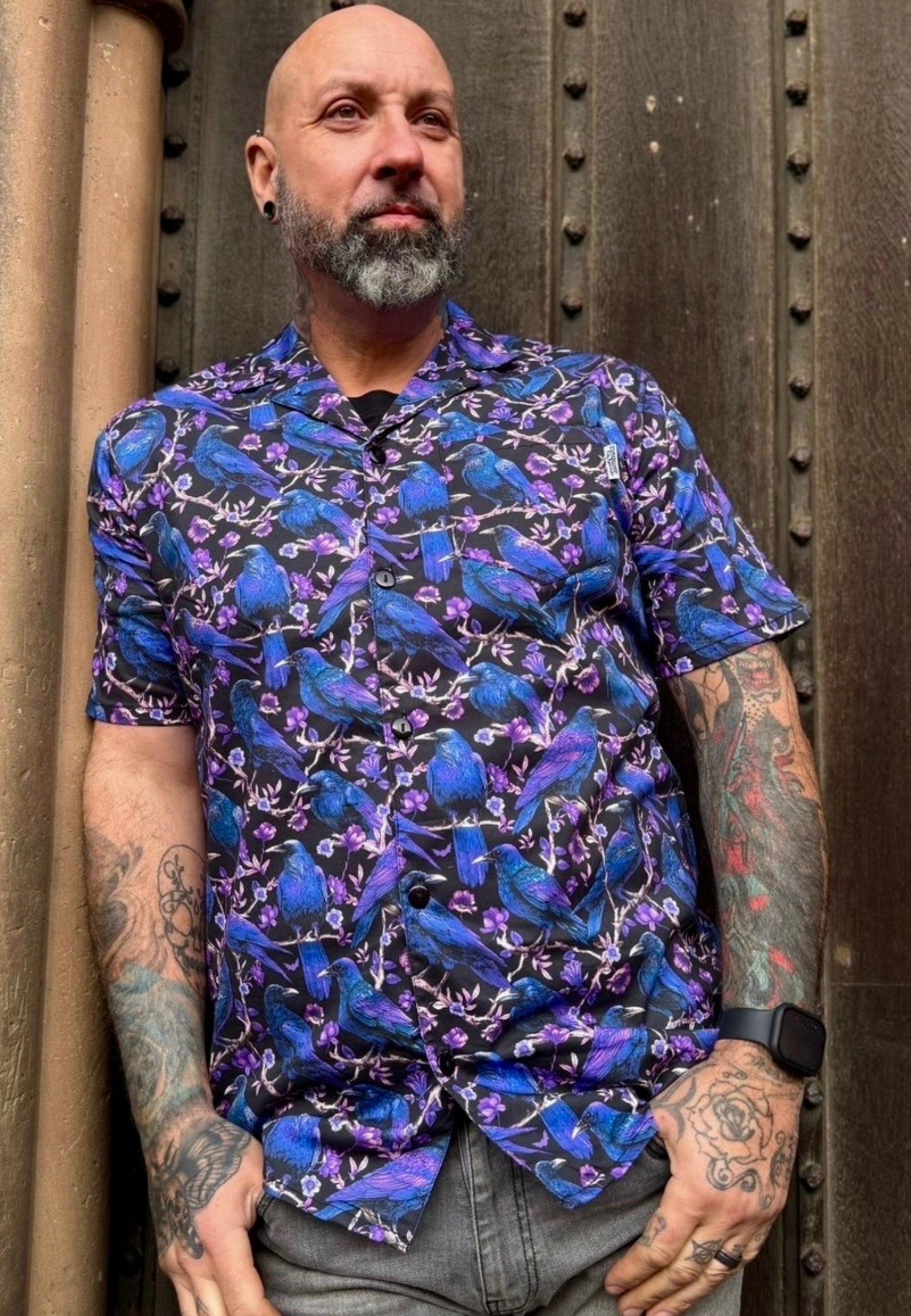 Man wearing a colorful shirt with a pattern of ravens and crows, standing against a textured wall.