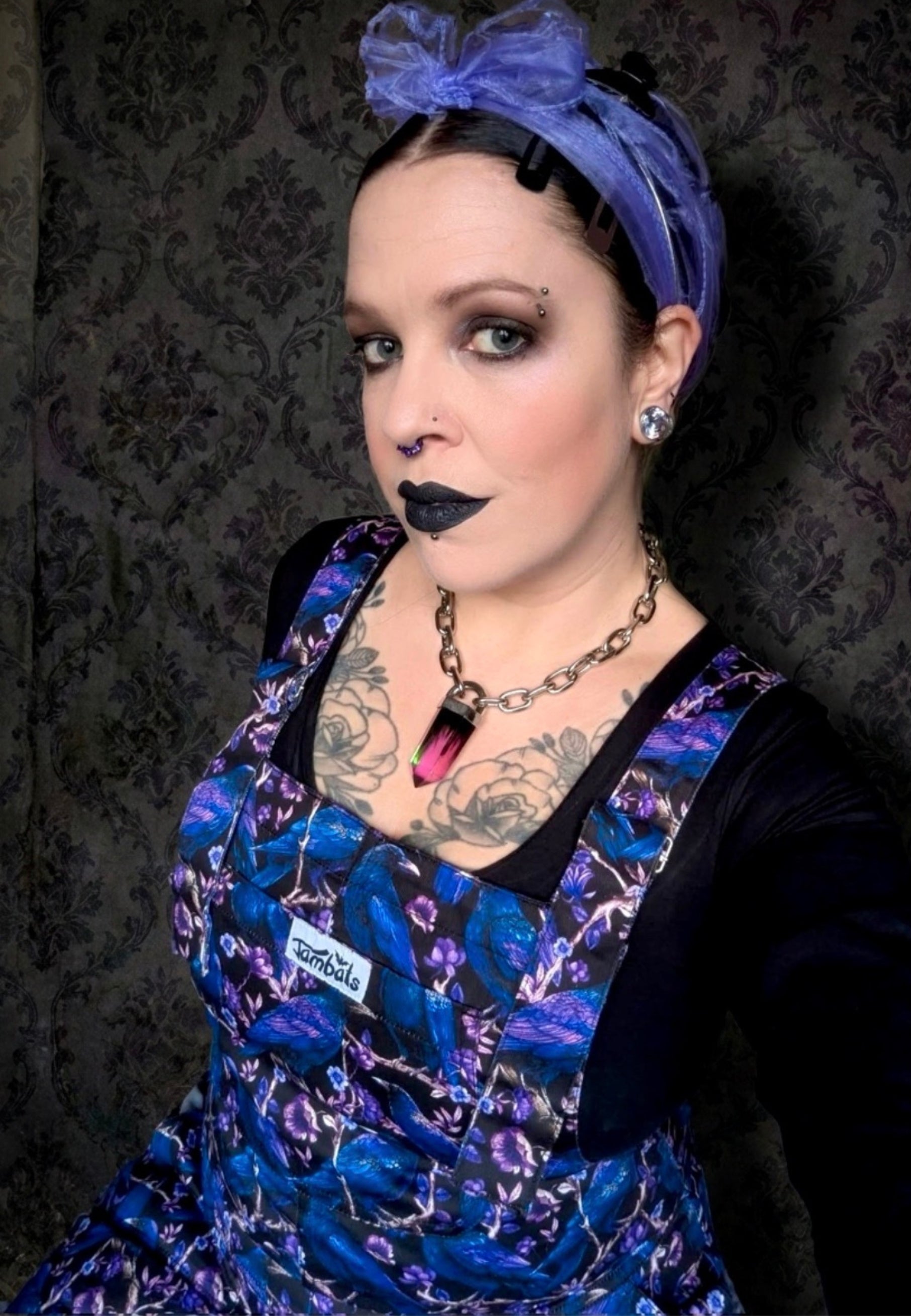 Close up of a woman wearing dungarees with a beautiful Corvid print. Deep purple and blue crows and raven on a black background. Woman is also wearing a black top and purple chiffon hair scarf. Background is vintage gothic style wallpaper. 