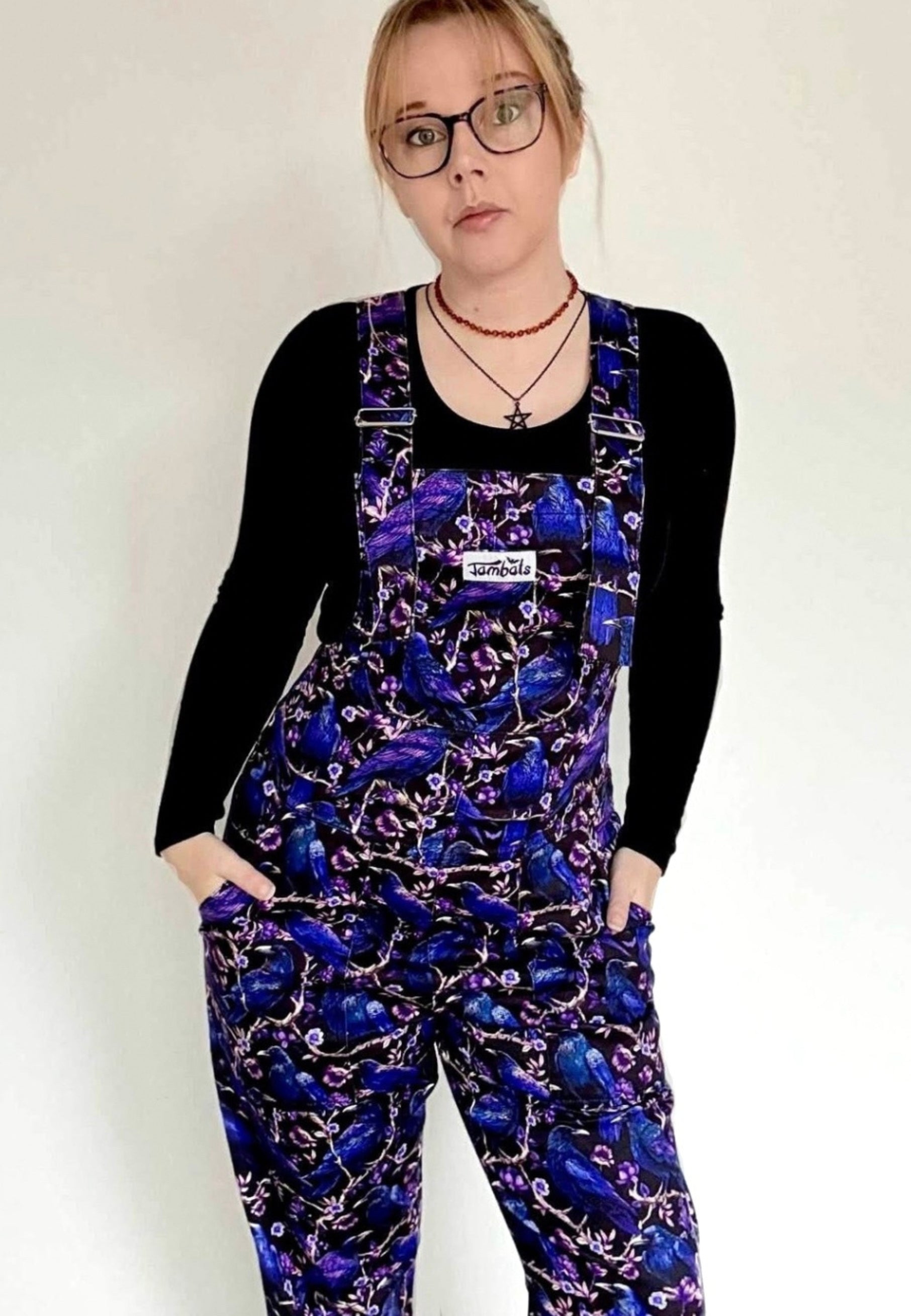 Woman wearing dungarees with a beautiful Corvid print. Deep purple and blue crows and raven on a black background. Woman is also wearing a black top and glasses. Studio photo