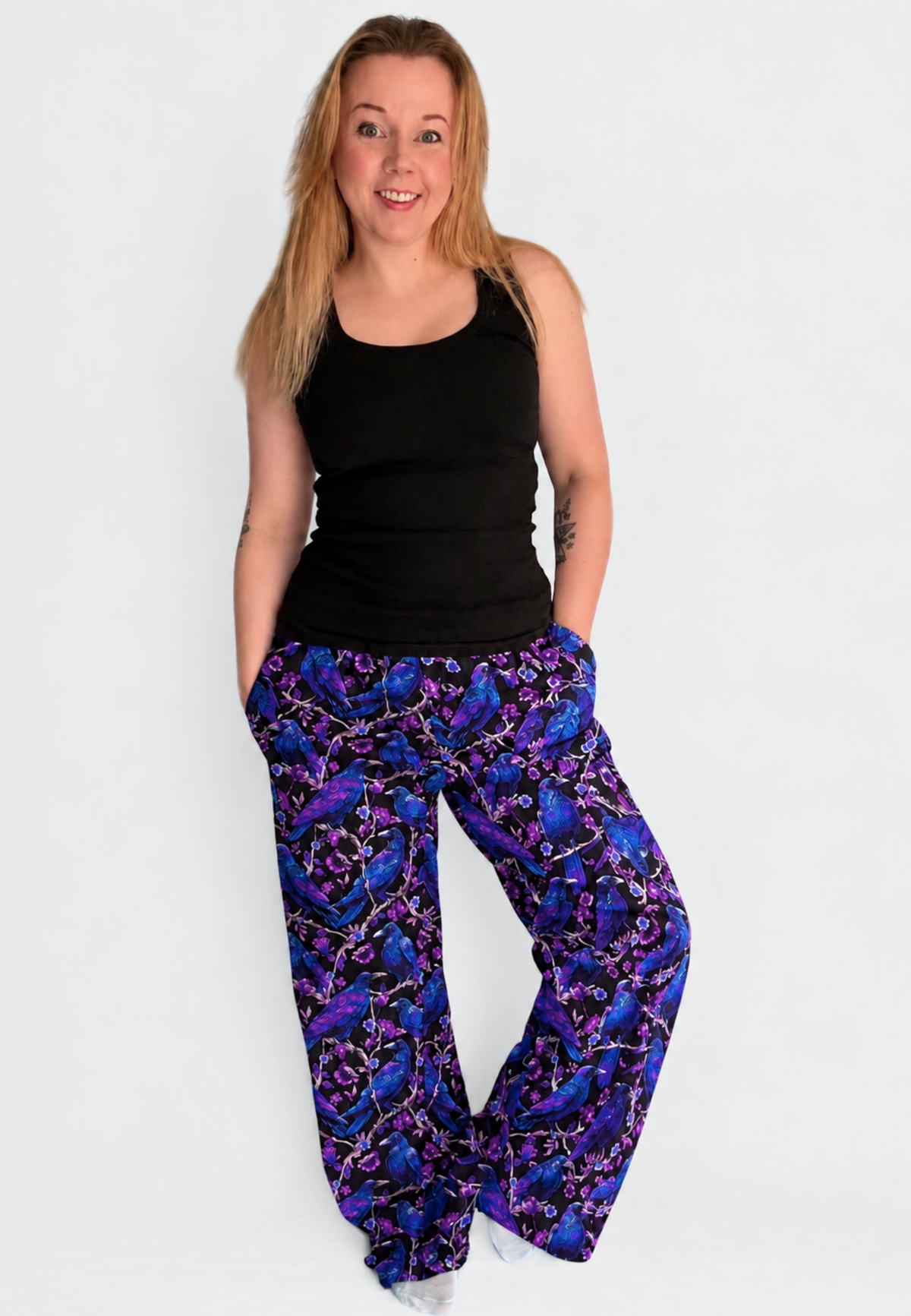 A woman modelling a pair of wide leg lounge pants with a vibrant purple and blue soft gothic raven and crow print , paired with a black tank top, against a plain light gray background.