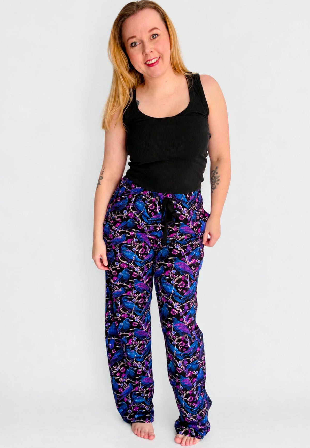 A woman modelling a pair of wide leg lounge pants with a vibrant purple and blue soft gothic raven and crow print , paired with a black tank top, against a plain light gray background