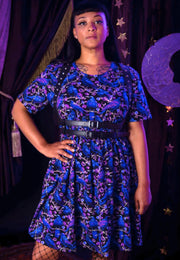 A woman modelling a black smock patterned dress with a black belt, standing against a dark curtain backdrop with decorative stars and a moon cutout. The dress has a beautiful print of cries and ravens on blossom trees in purples, blues and black 