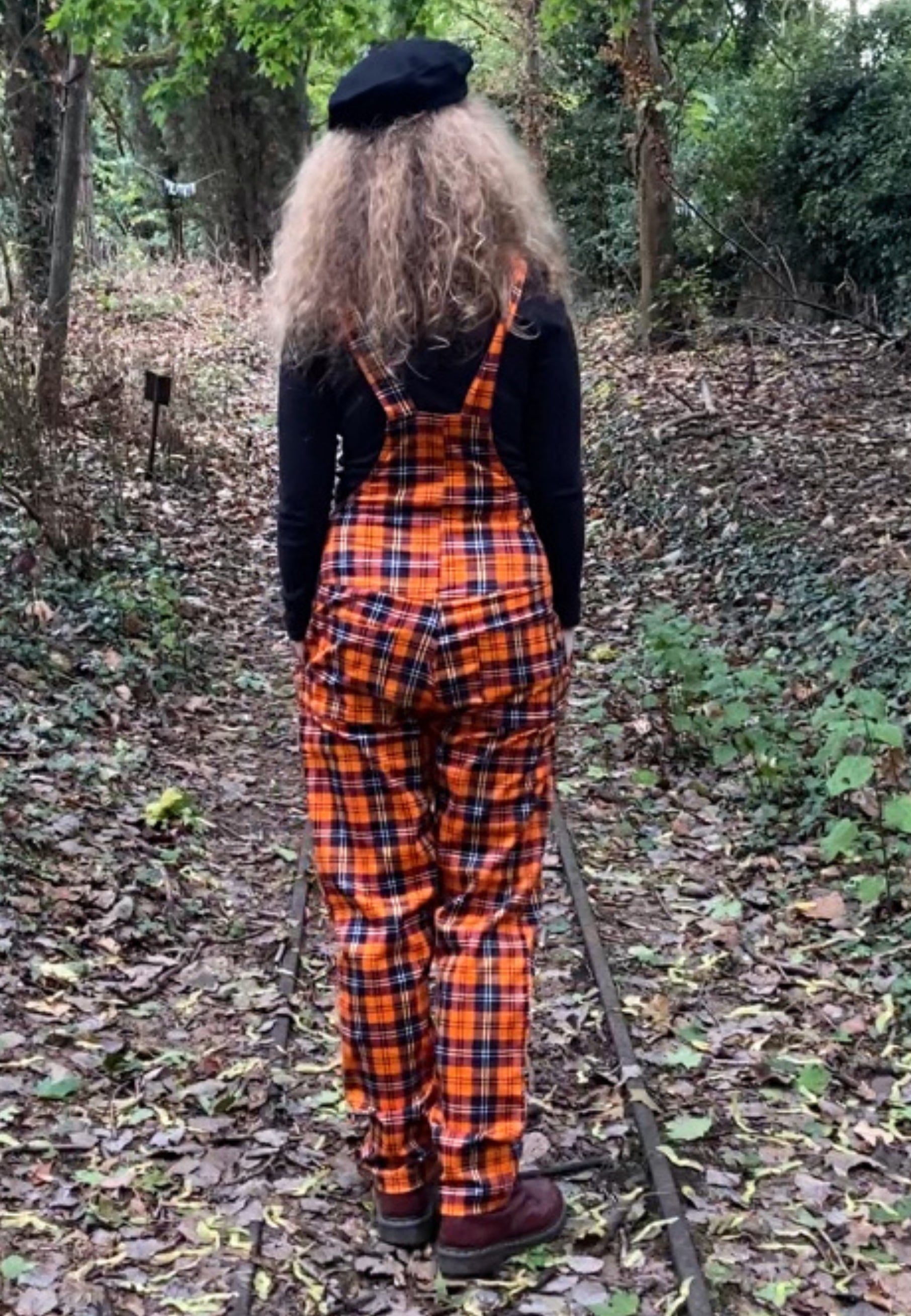 Woman wearing orange tartan dungarees overalls in a forest setting. View is showing the shape of the dungarees from the back