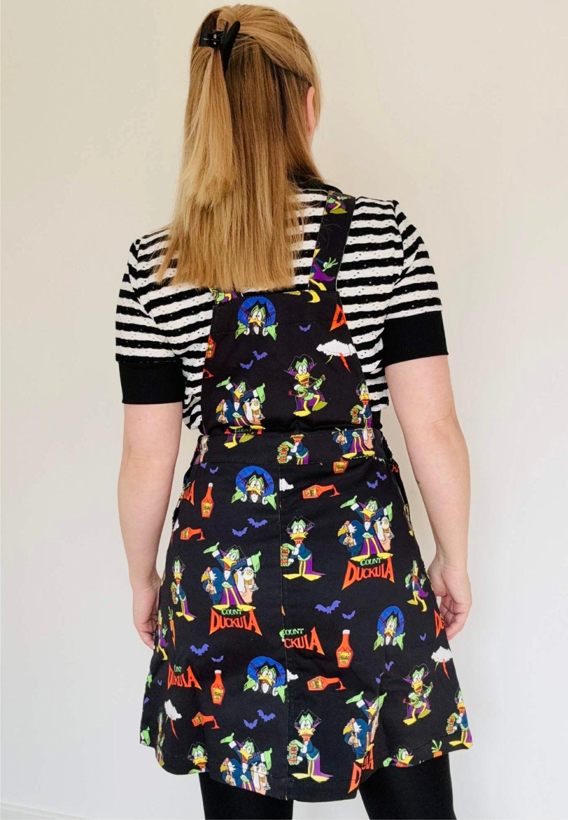 Back view of a woman wearing a black pinafore dress with vibrant Count Duckula print. Studio photo. 