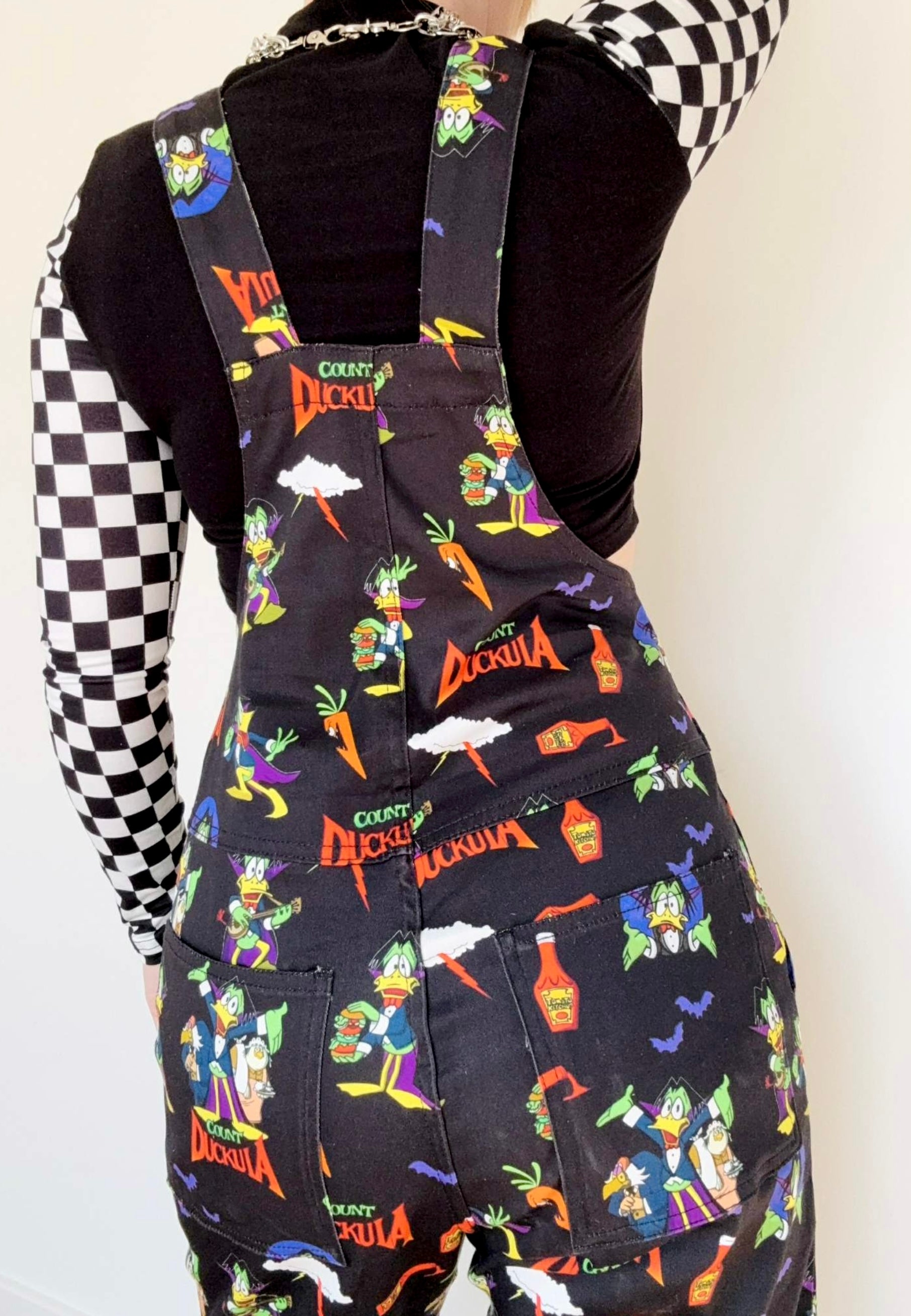 Back view of a woman wearing black Count Duckula print dungarees. Studio photo. 