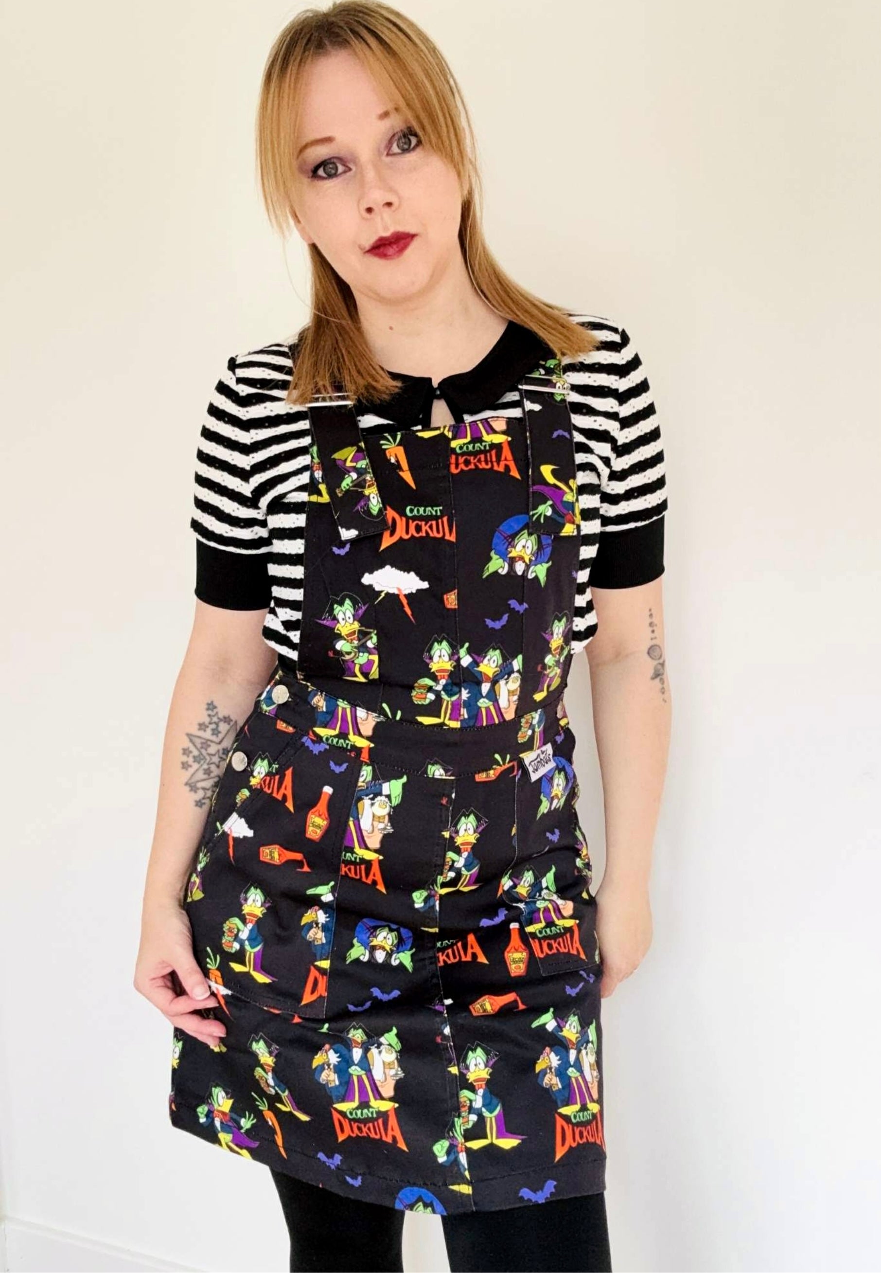 Woman wearing a black pinafore dress with vibrant Count Duckula print. Studio photo. 