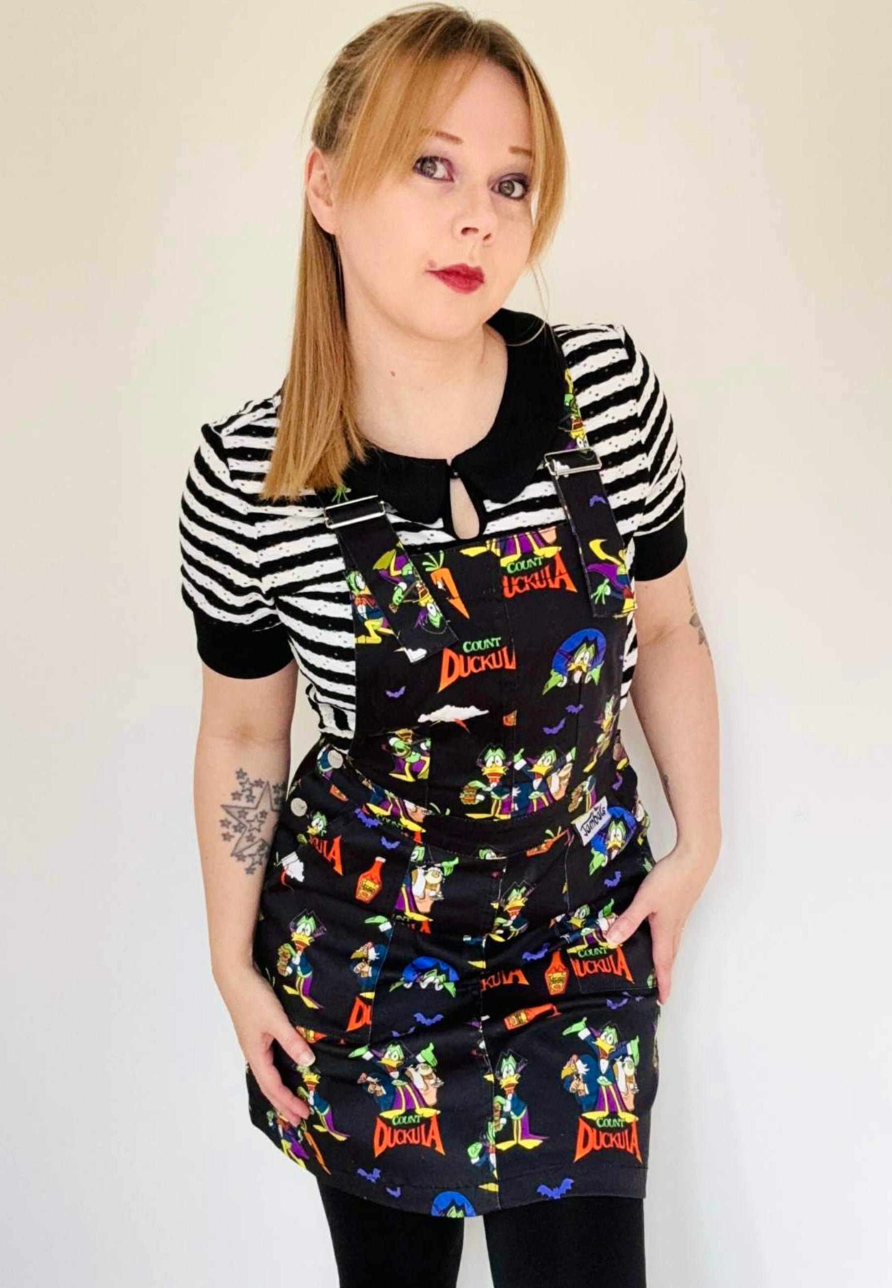 Woman wearing a black pinafore dress with vibrant Count Duckula print. Studio photo. 