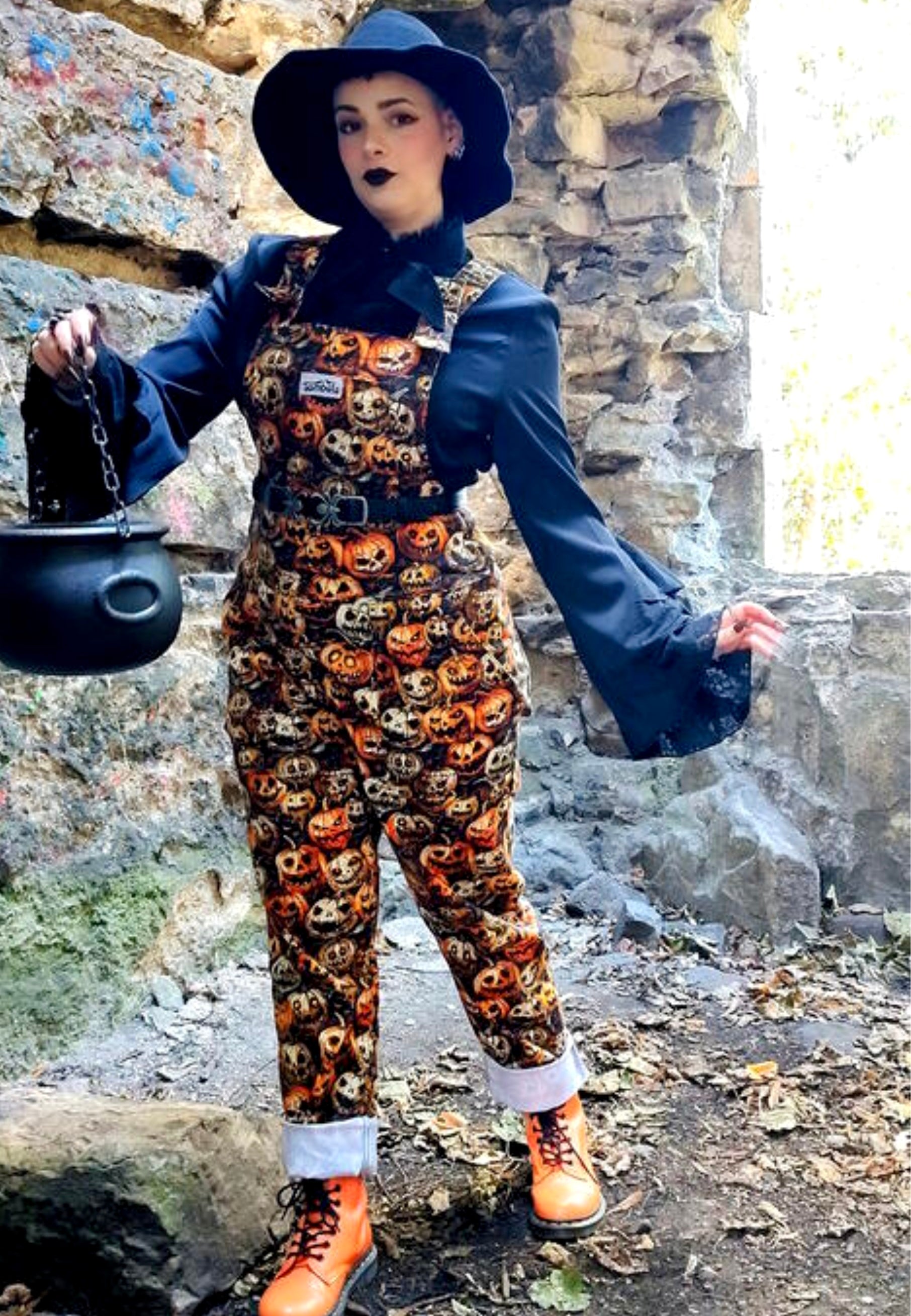 Model wearing a witches hat and Halloween themed dungarees overalls with scary creepy pumpkins print , also wearing orange boots in a spooky urban outdoor setting. Holding a cauldron. Creepy Gothic Halloween aesthetic