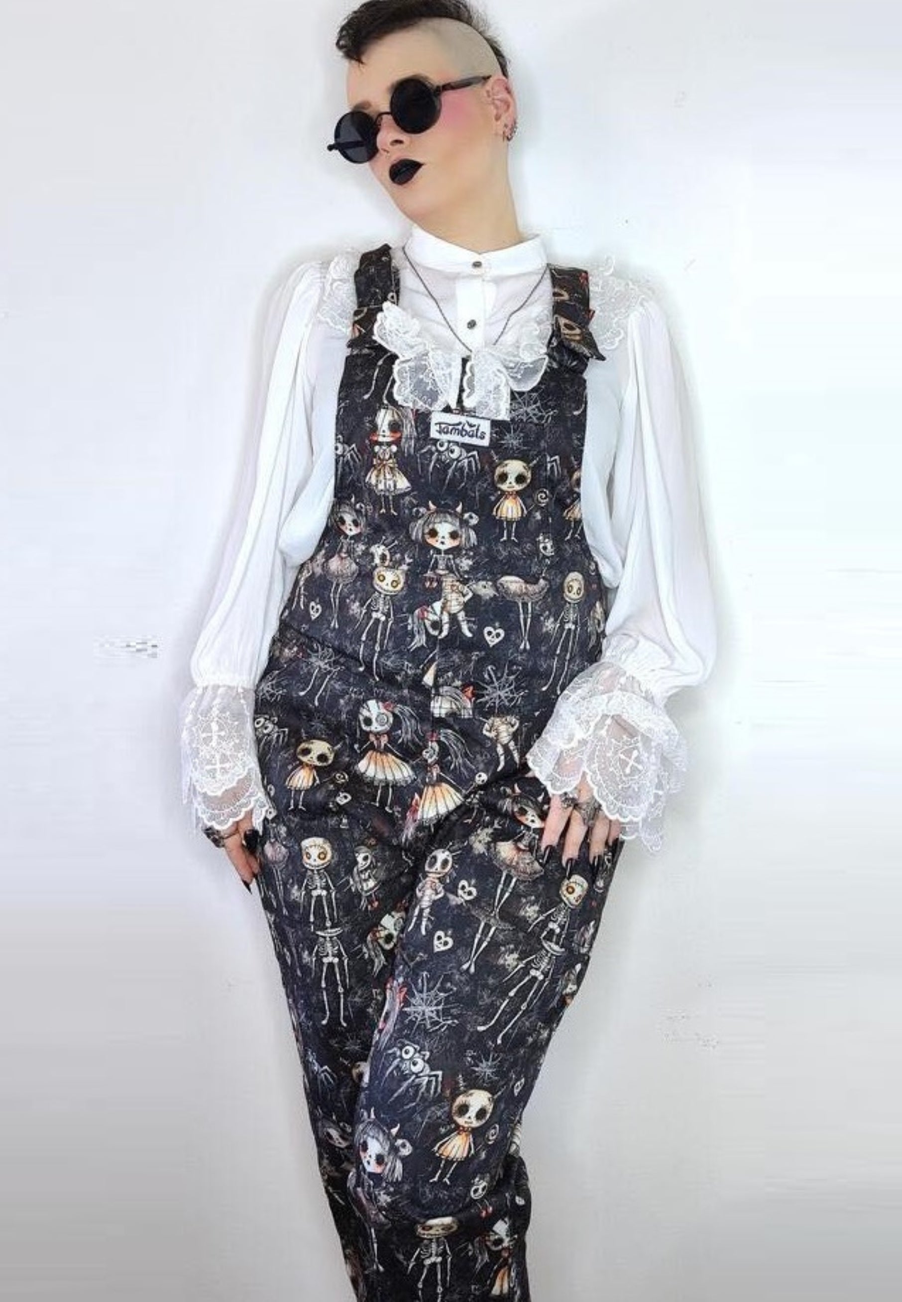 Model wearing dungarees with a pattern of cute but creepy haunted dolls in various poses against a dusty background. Model is also wearing a white frilly sleeved shirt and sunglasses. Alternative Goth style. Jambats dungarees.  