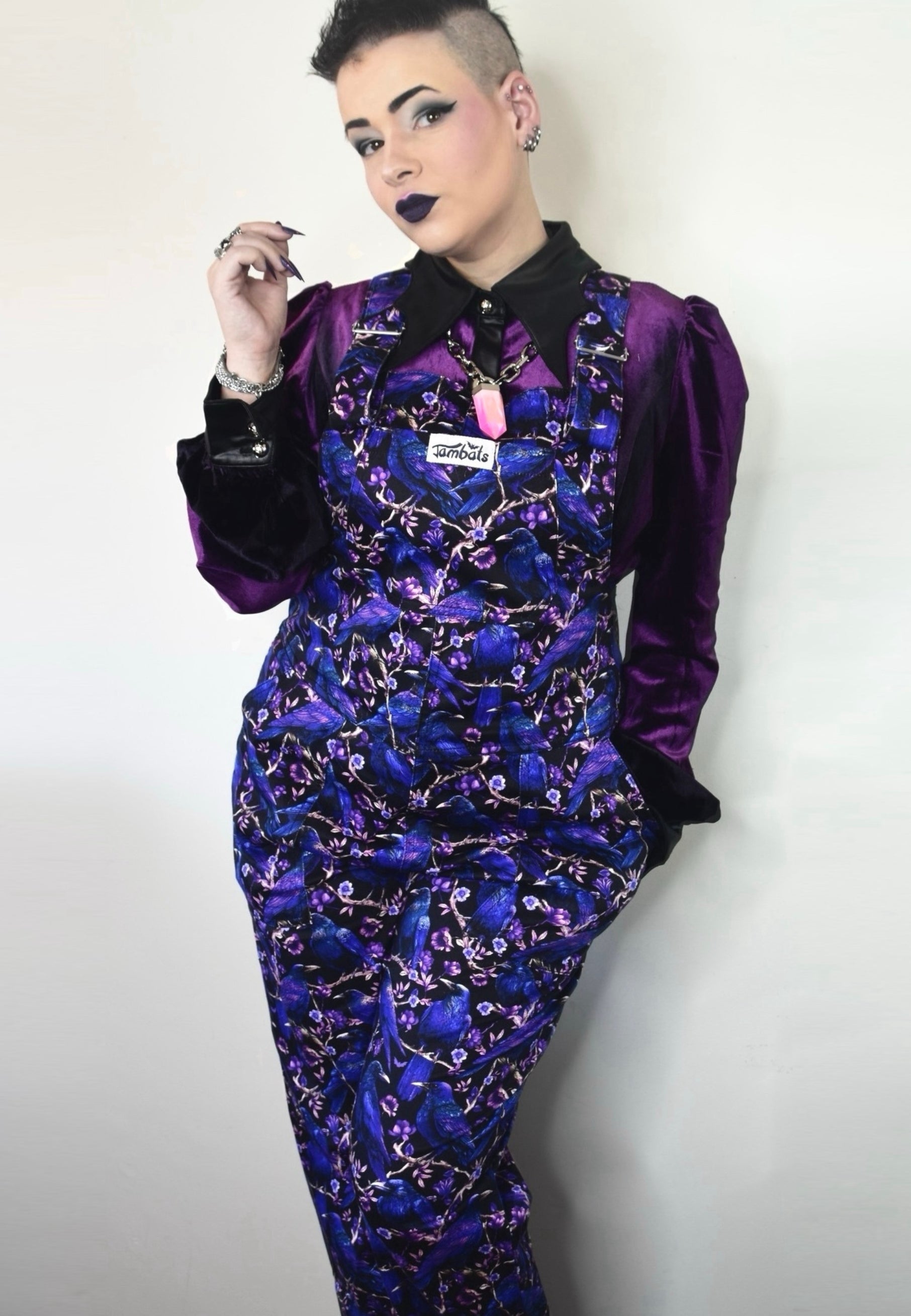 Woman wearing dungarees with a beautiful Corvid print. Deep purple and blue crows and raven on a black background. Woman is also wearing a purple velvet blouse. Studio photo