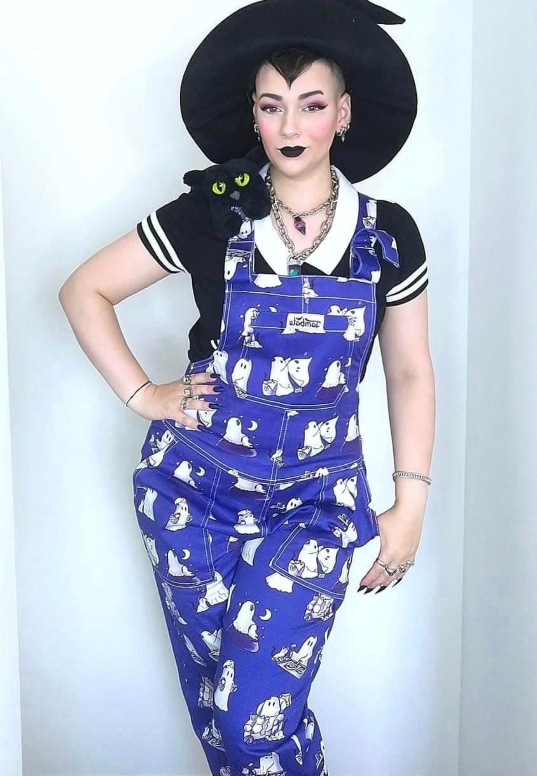 A witchy woman in a witches hat wearing cute blue dungarees with a print of ghosts and ghost cats. 