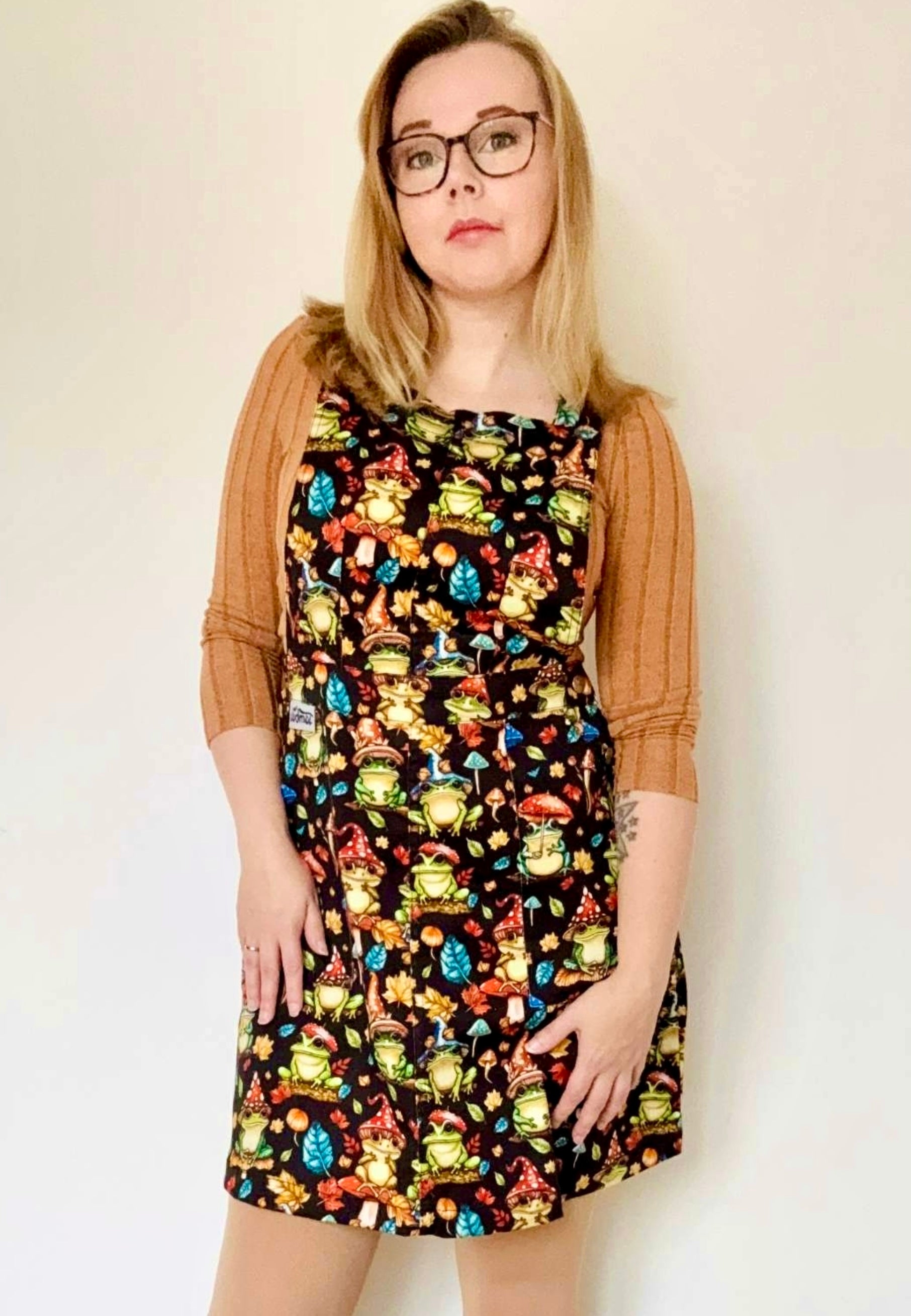 Woman in bookish glasses wearing a pinafore dress with a whimsical print of frogs with toadstool hats and toadstool umbrellas. Studio photo. 