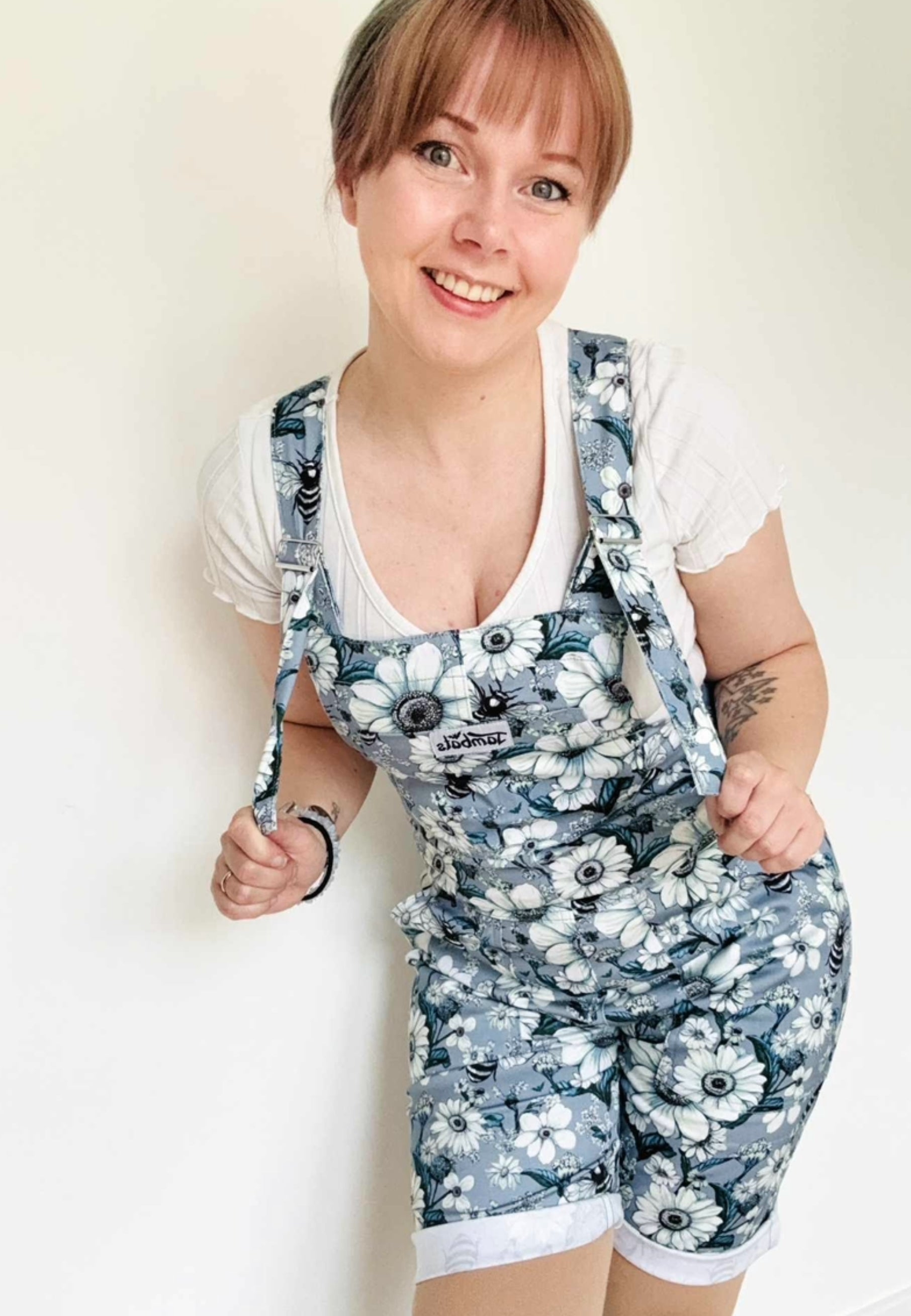 Woman wearing dungaree overall shorts with a print of daisies and bees and a white shirt underneath on a plain background. Overalls  dungarees shorts. 