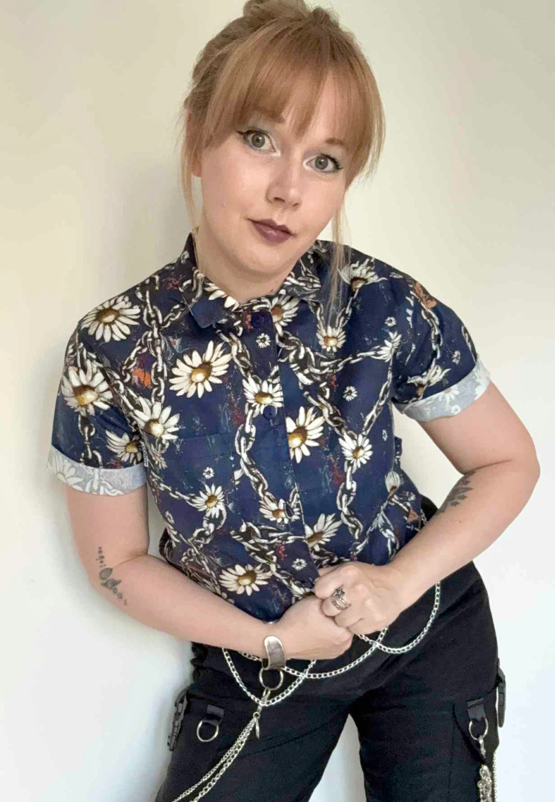 Woman wearing a deep blue shirt  with chains and daisies and dark pants against a plain background. Grungey floral style print. 