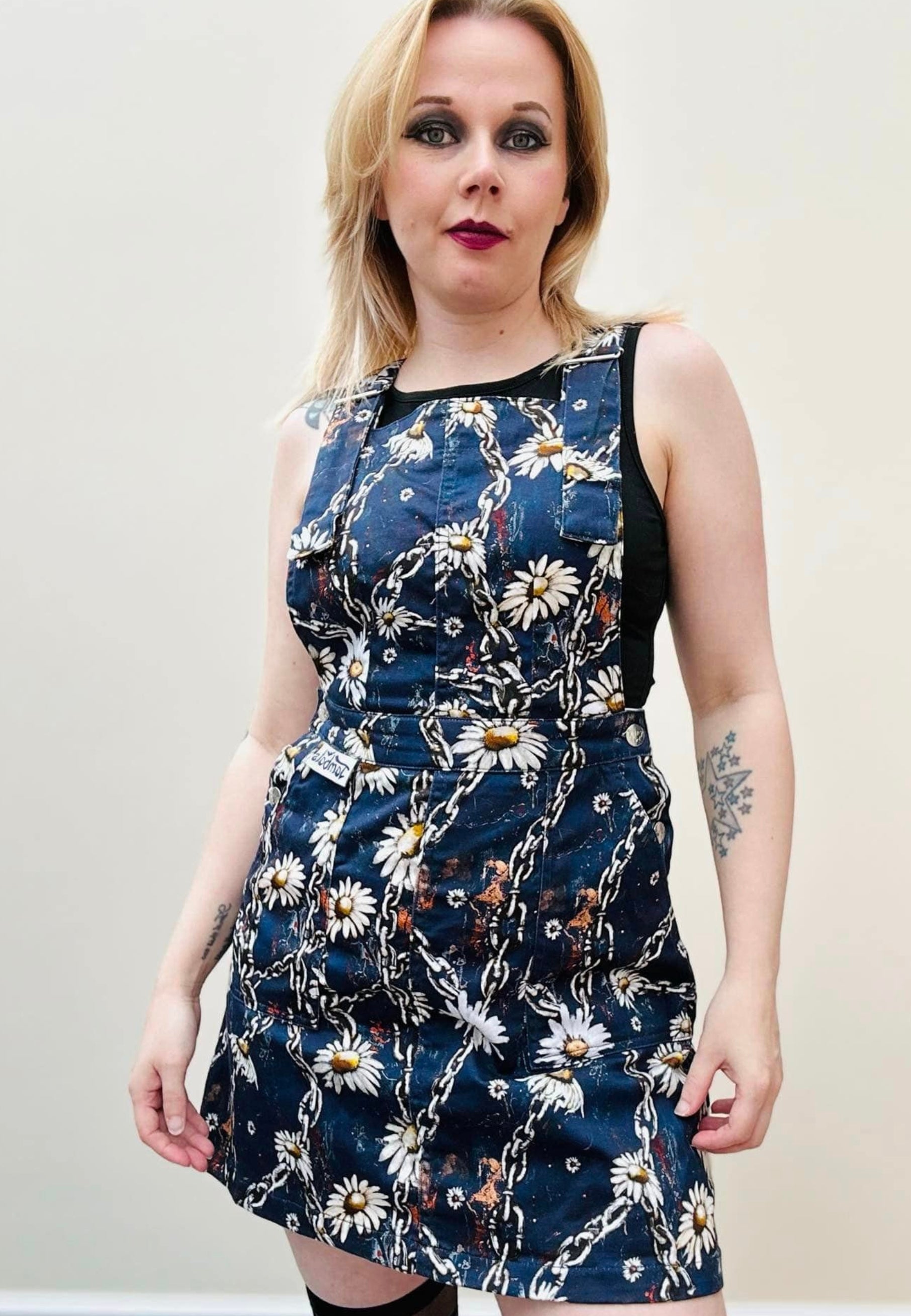 A woman wearing a deep blue pinafore dress with a unique urban grunge Daisy Chains print, featuring deep hip pockets and side button fastenings. Alternative styling.
