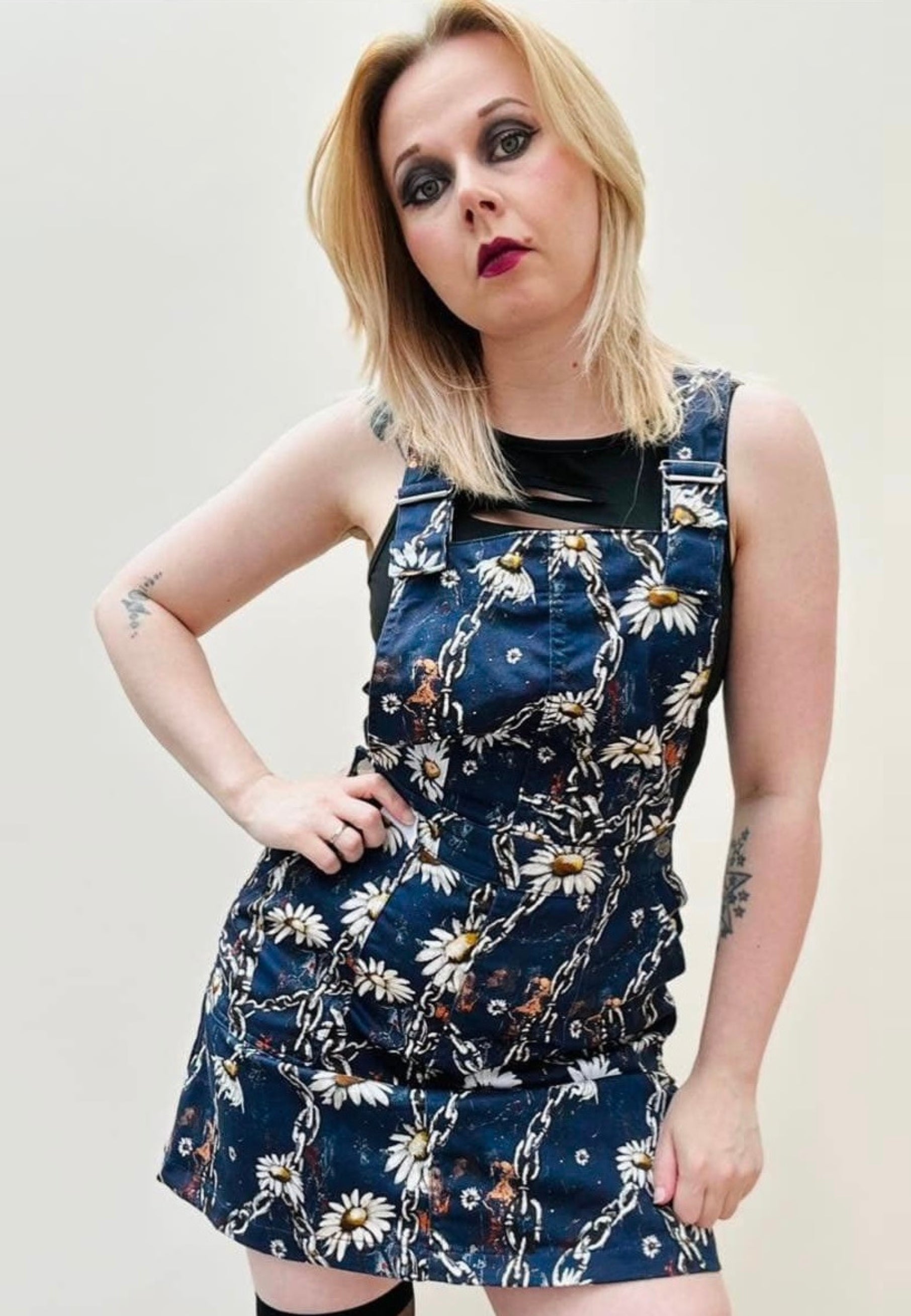 A woman wearing a deep blue pinafore dress with an urban grunge Daisy Chains print, featuring deep hip pockets and side button fastenings. Alternative styling.