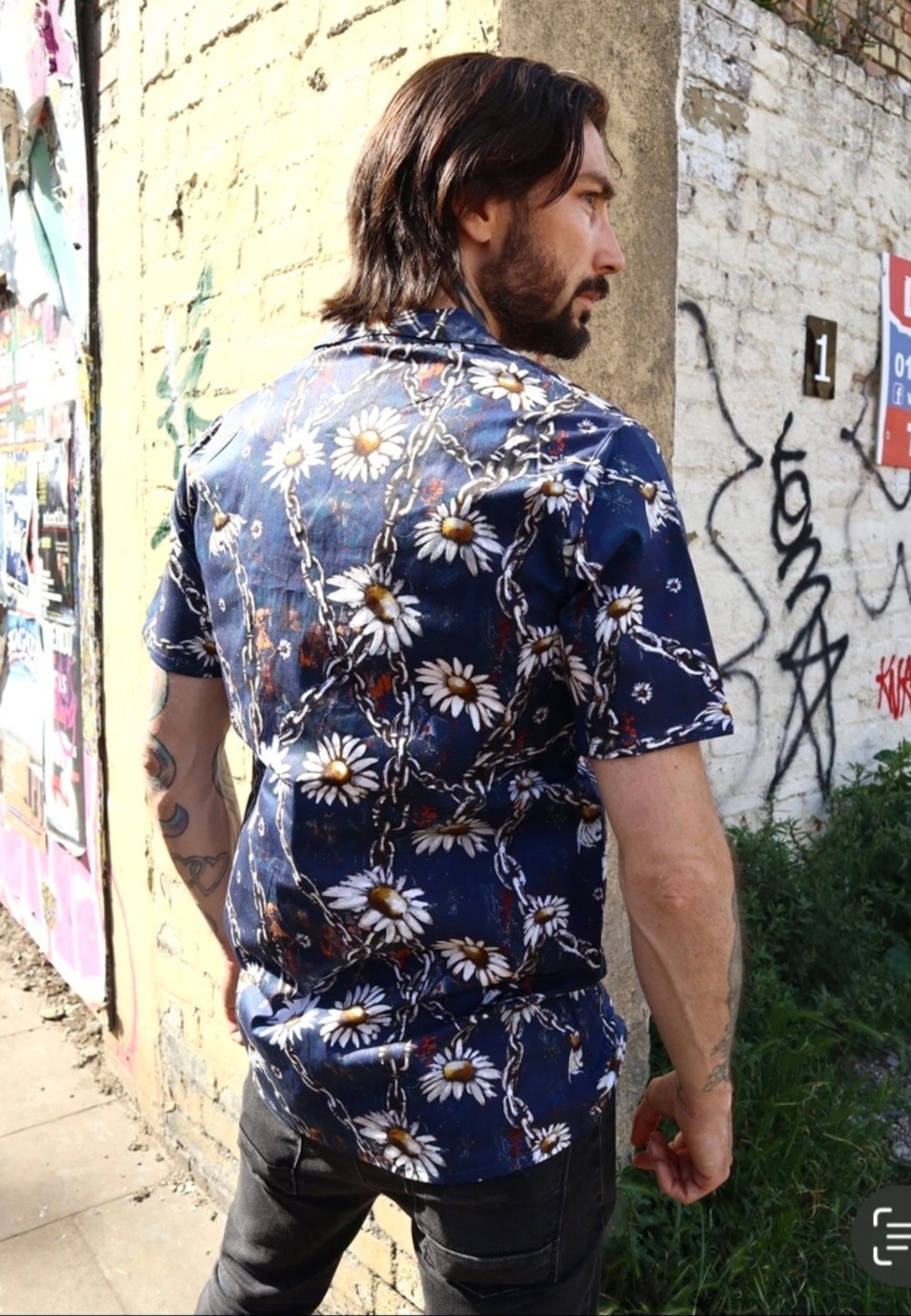 Man wearing a blue shirt with a daisy and urban grungey chains prong against a graffiti covered wall. 