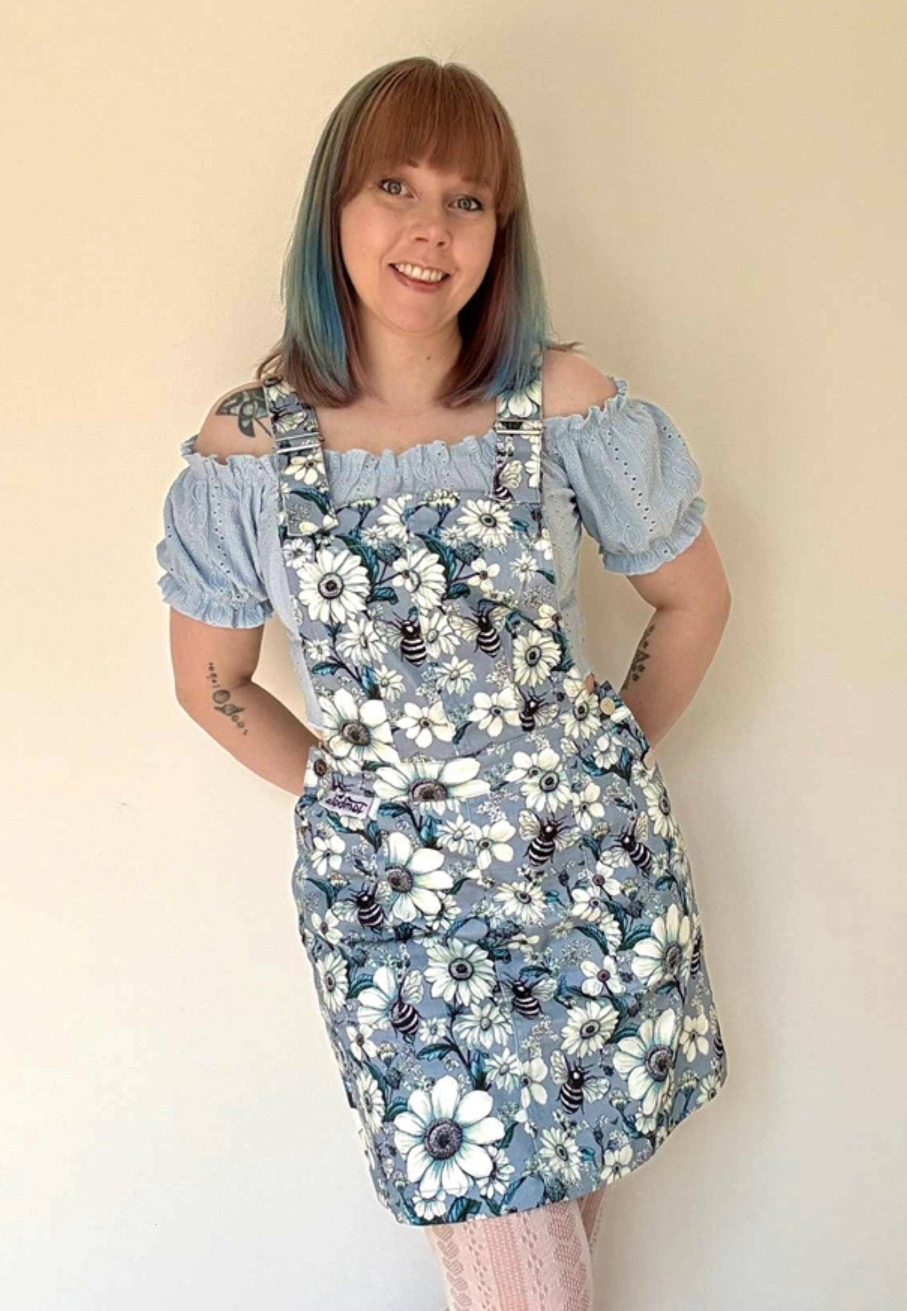 Woman wearing a floral pinafore dress with a print of daisies and bees,over a blue off-shoulder top against a beige background