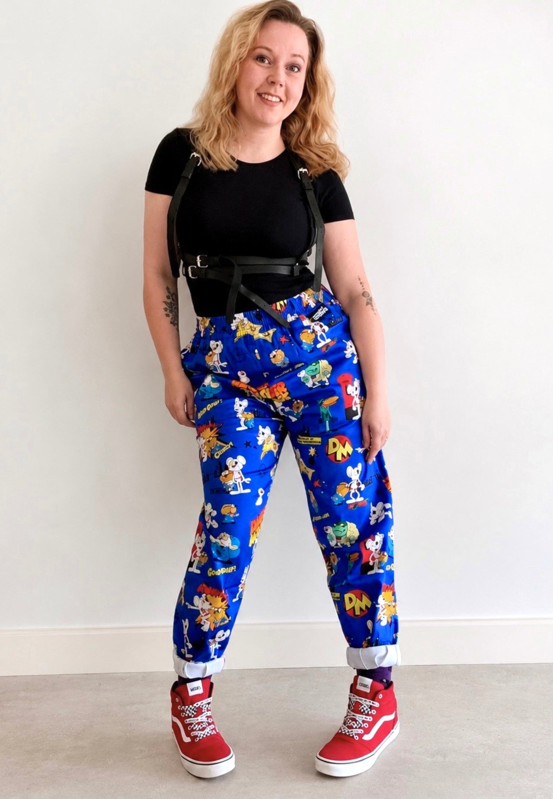 A woman modeling a pair of bright blue trousers with a colorful character print of the 80’s icon Danger Mouse. Styled with a black t-shirt, black harness, and red Vans against a plain white wall.