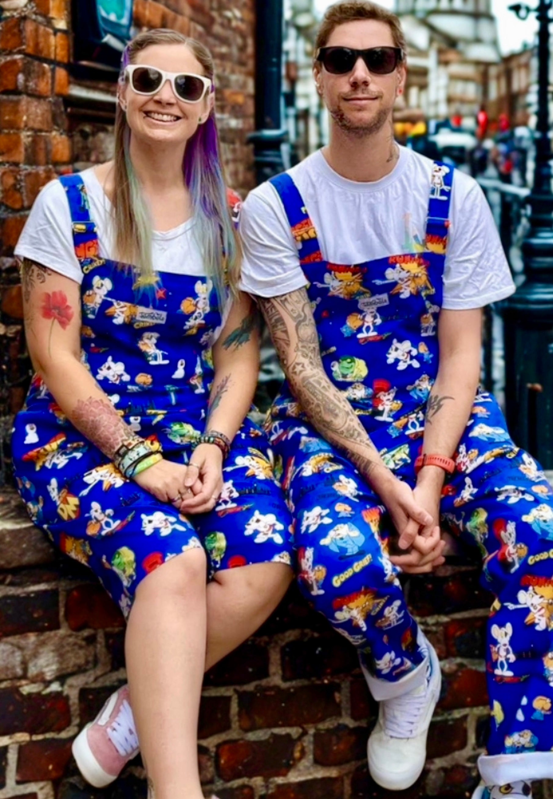 Two people wearing matching blue dungarees with a Danger Mouse pattern  sitting on a brick wall.