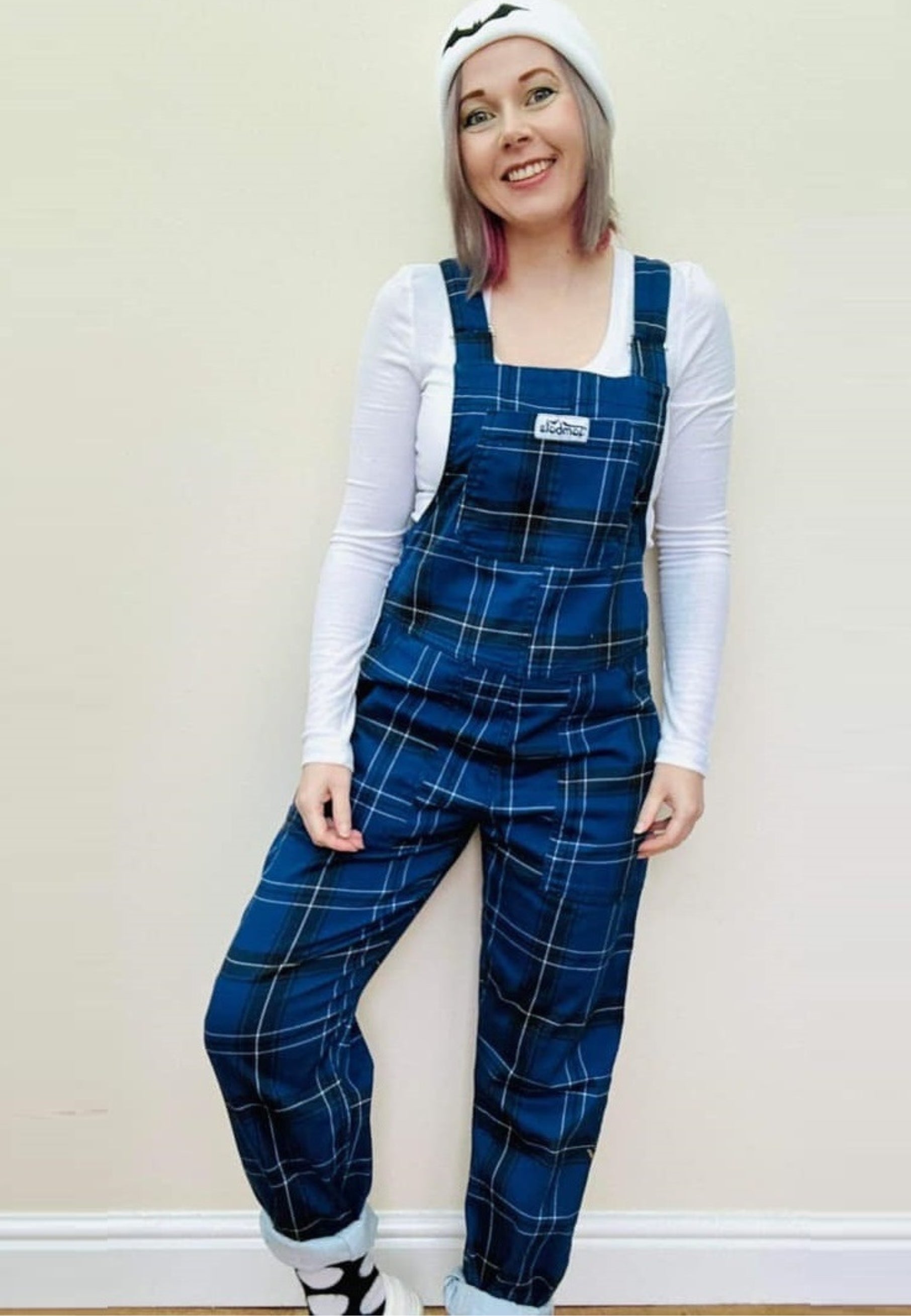 Model in classic dark blue and black tartan dungarees, white top and white beanie hat. Studio photo on a plain background