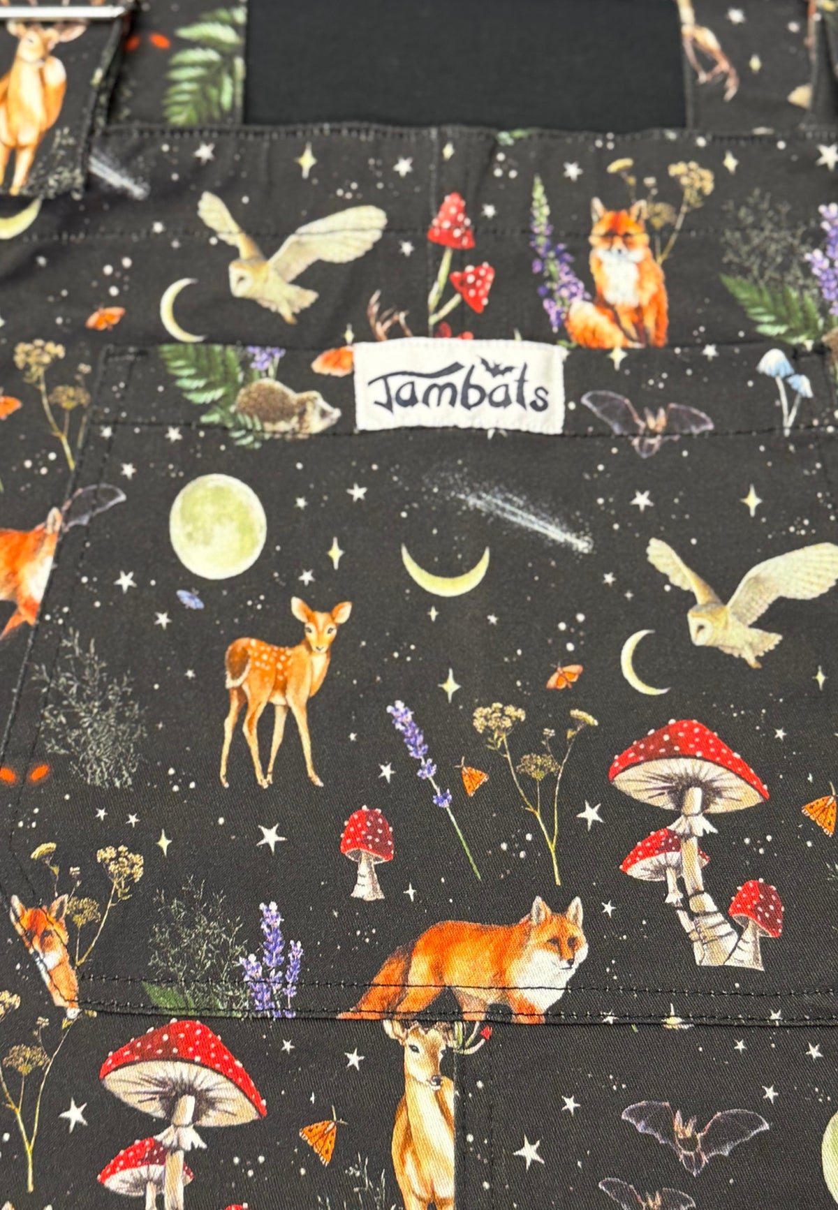 A close up of a black fabric dungarees  featuring a whimsical woodland print with foxes, deer, mushrooms, owls, stars, and moons. A white fabric label reading 'Jambats' is stitched into the design.