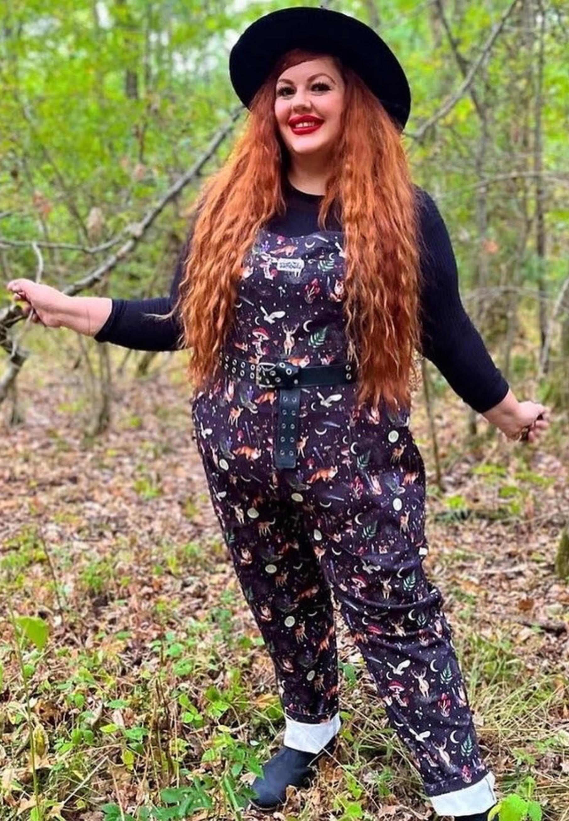 Plus size model in the woods wearing Jambats Dark Forest dungarees. Black print with  moonlit forest, stars, shooting stars, moons and night dwelling creatures like owls, foxes, deer. Beautiful overalls