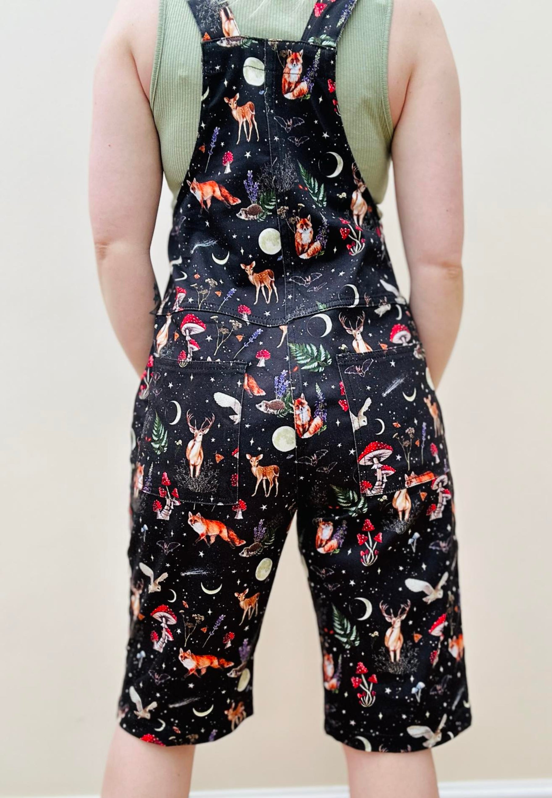 A woman wearing dark forest print shorts dungarees with a moonlit, star studded fabric and whimsical night creatures like foxes, deers, hedgehogs, owls, moon moths, and bats. Shorts dungarees, overalls. View from the back showing the back shape of the shorts overalls. 