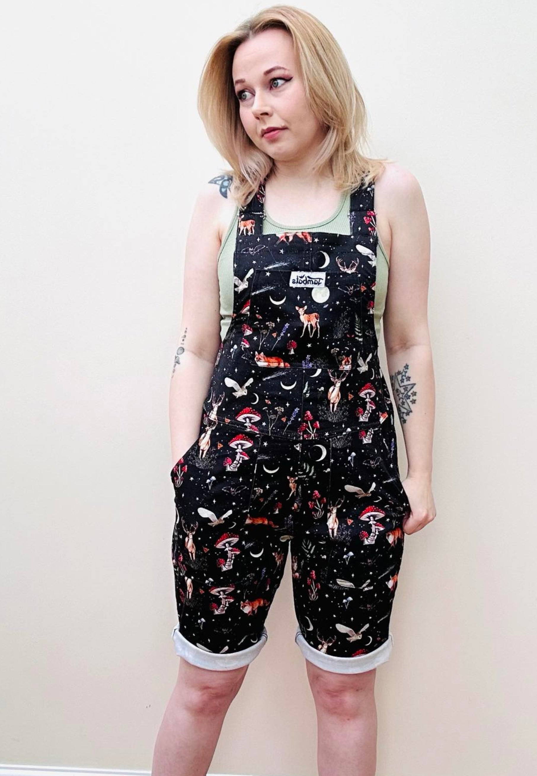 A woman wearing dark forest print shorts dungarees with a moonlit, star studded fabric and whimsical night creatures like foxes, deers, hedgehogs, owls, moon moths, and bats. Shorts dungarees, overalls. Cottagecore style. 