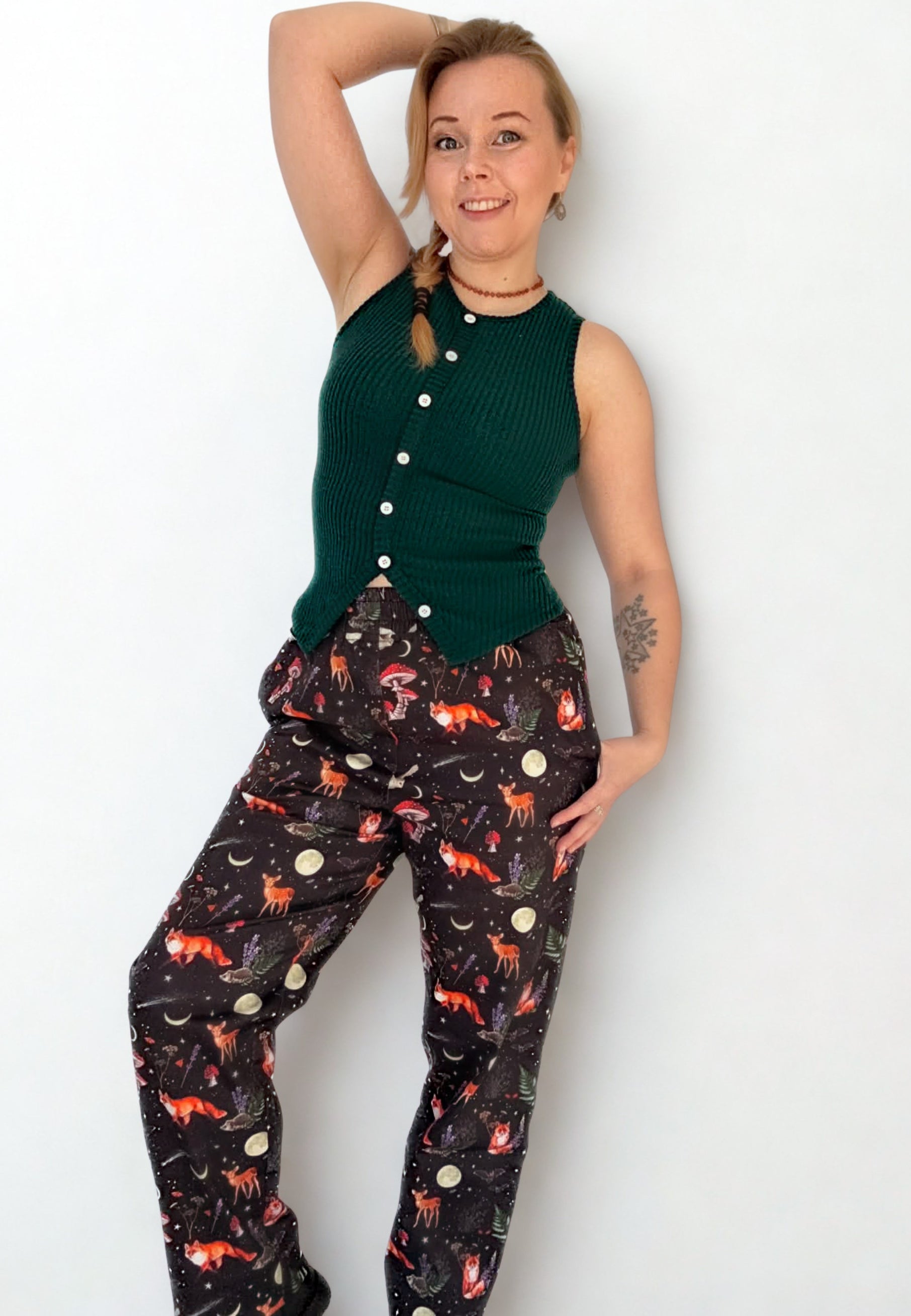 Woman with her hair in a plait wearing a green sleeveless top and black trousers with a beautiful whimsical Dark Forest Print. On a white wall, studio photo. 