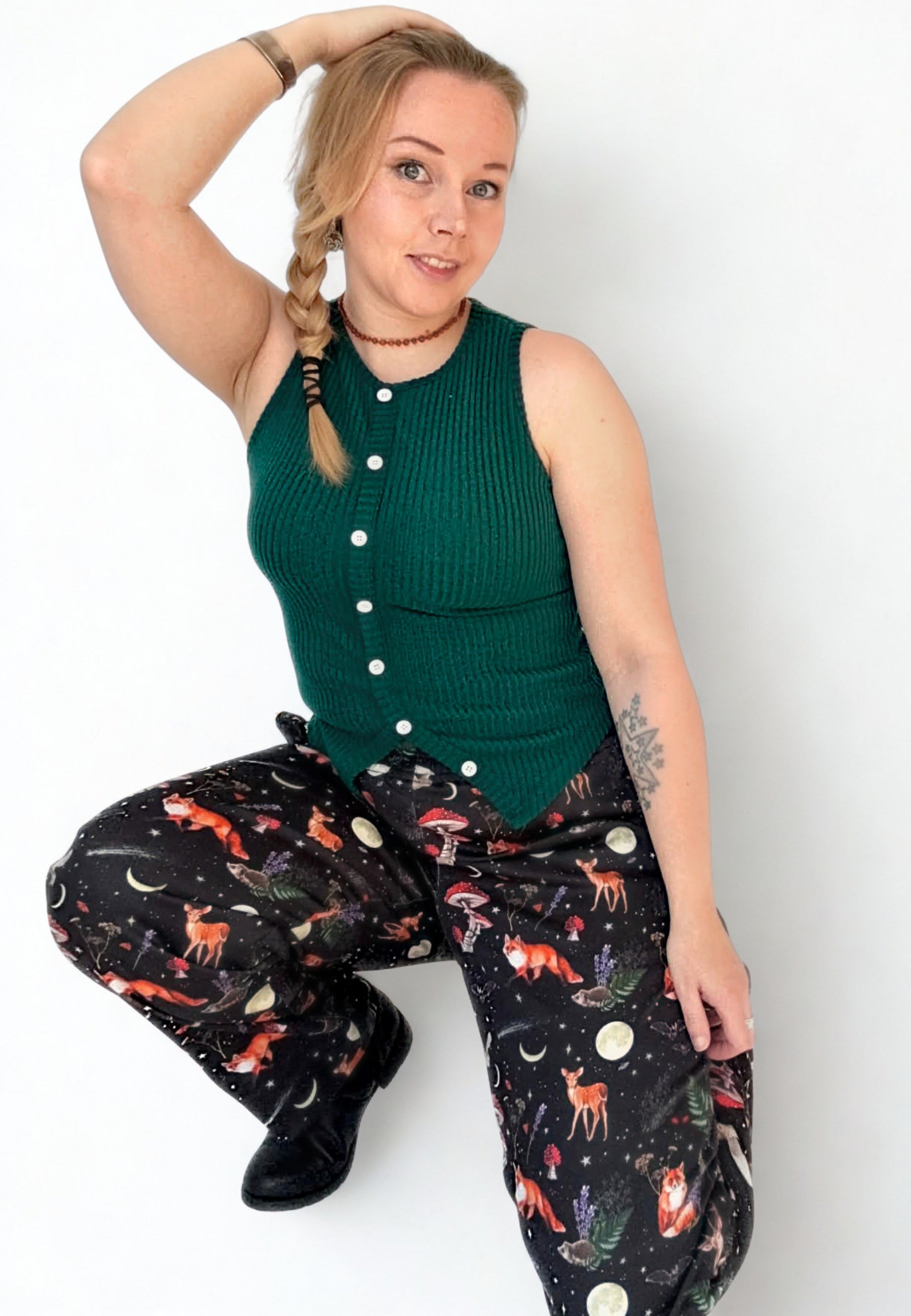 Woman with her hair in a plait wearing a green sleeveless top and black trousers with a beautiful whimsical Dark Forest Print. On a white wall, studio photo. Woman is crouching down 
