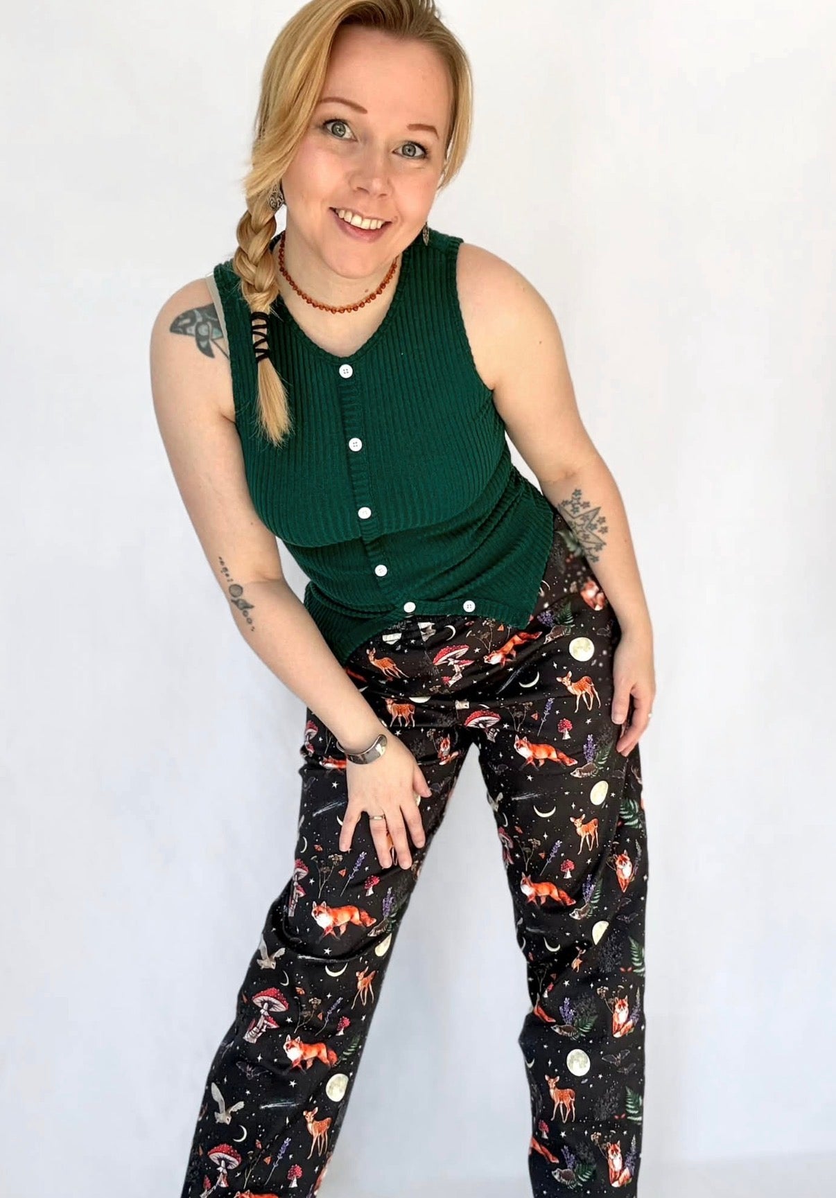 Woman with her hair in a plait wearing a green sleeveless top and black trousers with a beautiful whimsical Dark Forest Print. On a white wall, studio photo. 