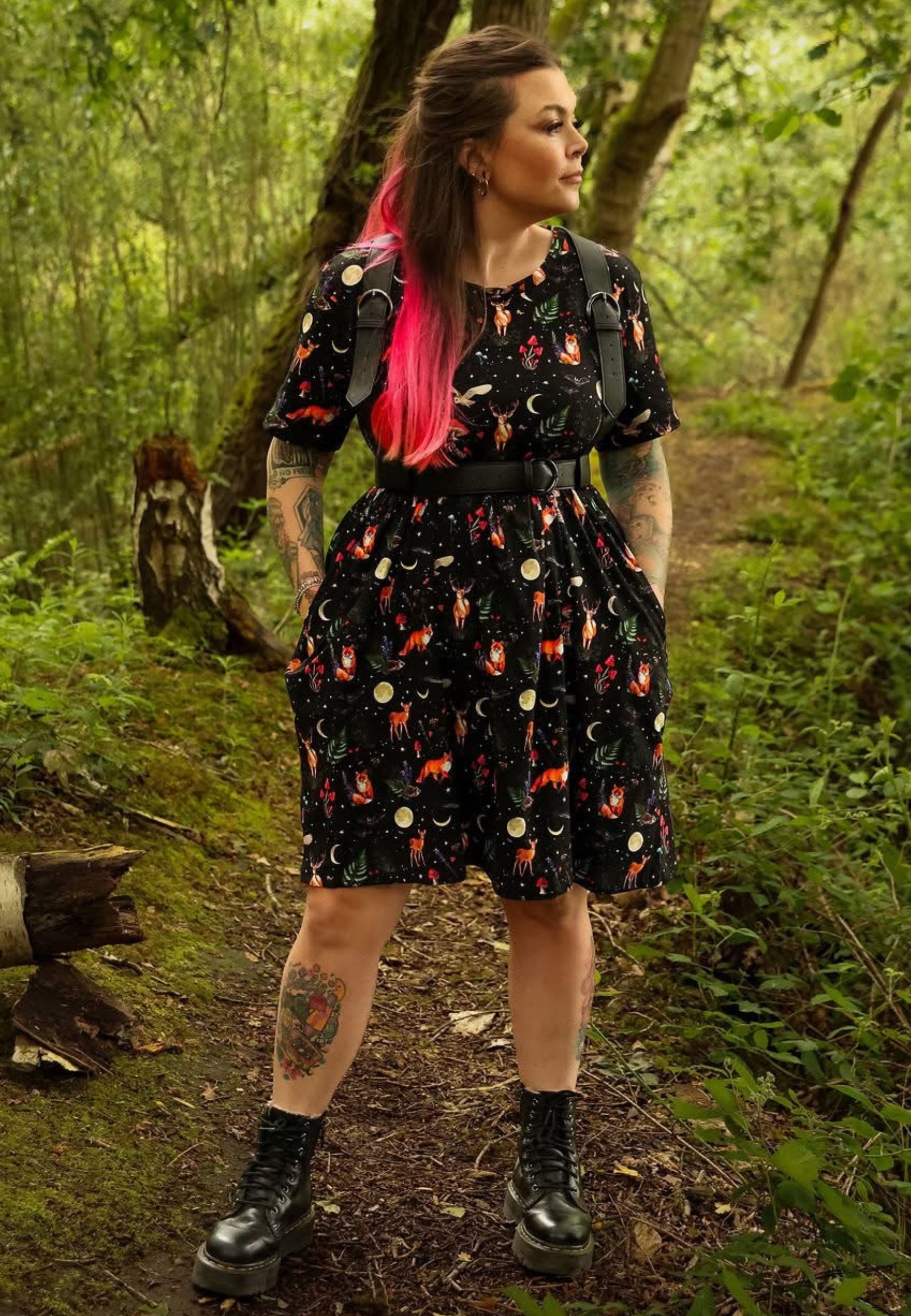 A woman stands on a forest path wearing a black dress with a whimsical print of foxes, moons, and stars, along with black boots 
