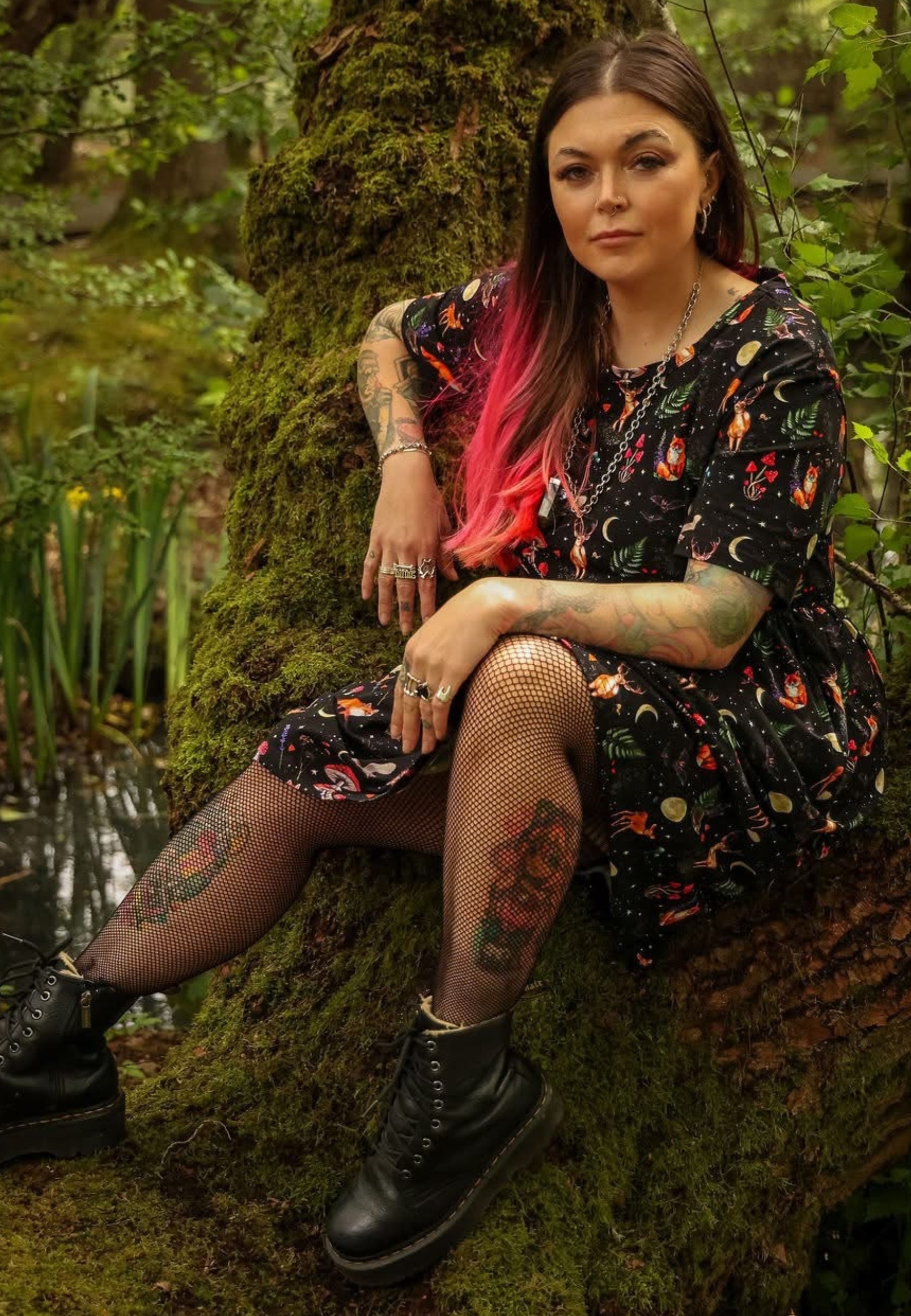 A woman with pink-tipped hair sits on a mossy tree trunk in a forest, wearing a black dark whimsical woodland print dress, fishnet tights, and black boots.