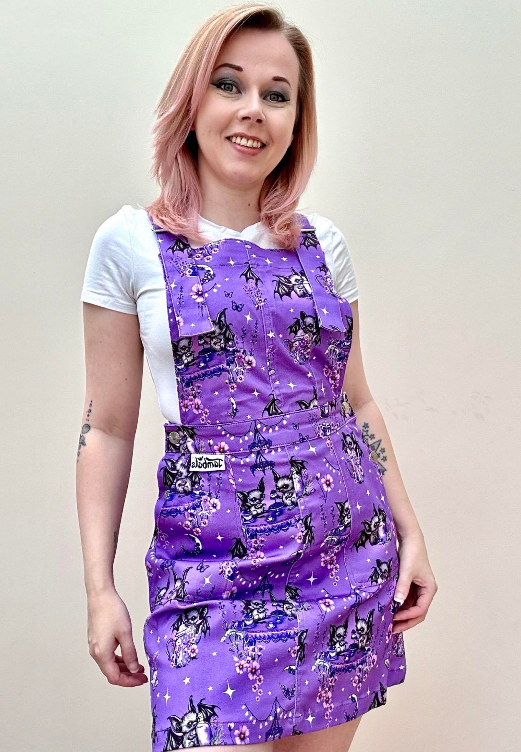 A woman wearing a deep lavender purple pinafore dress with a cute and whimsical bats tea party print, over a white t-shirt, standing against a neutral background. Cute goth print pinafore dress. 