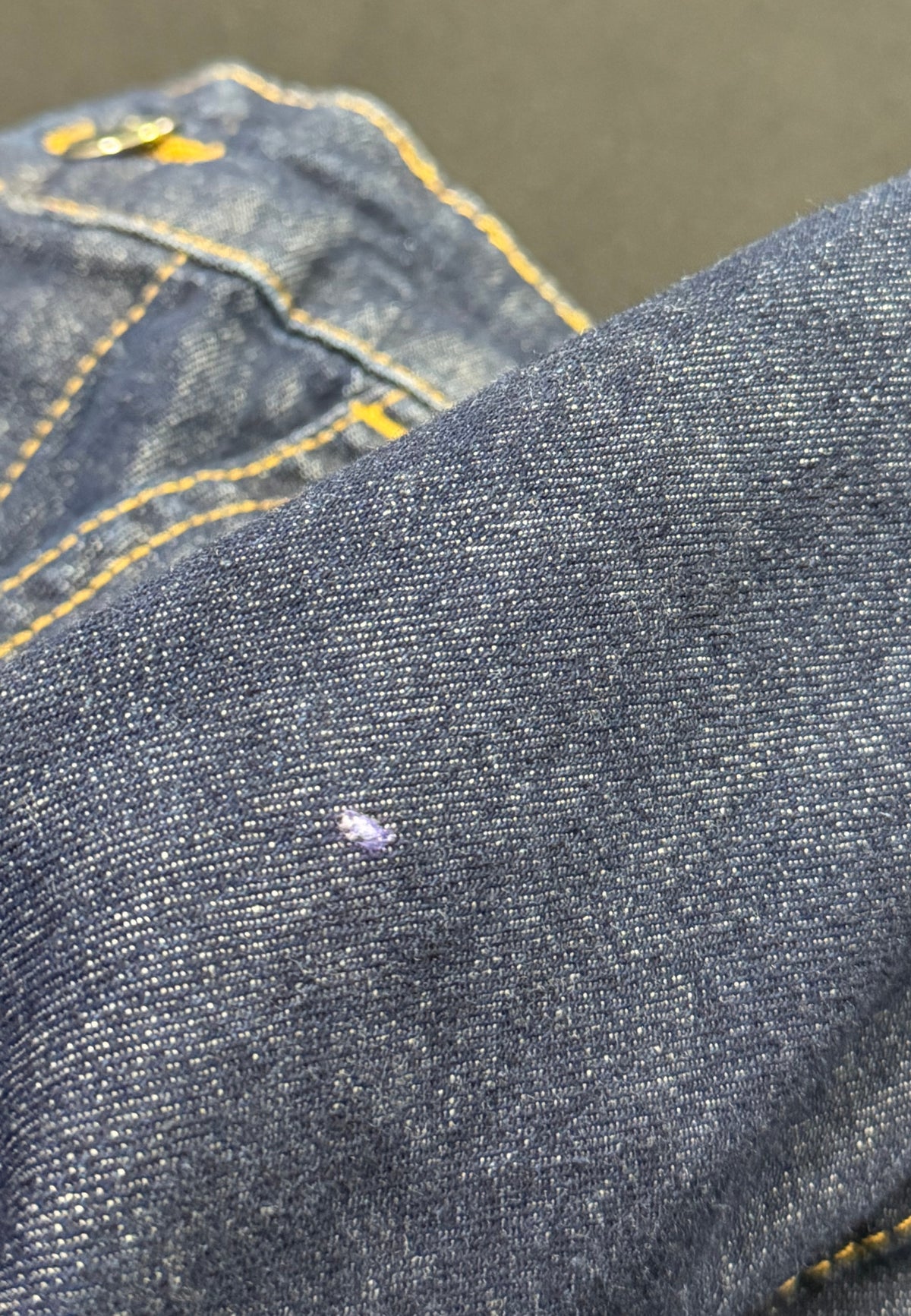Close up of dark blue denim fabric with yellow contrast stitching and a small hole on the front.