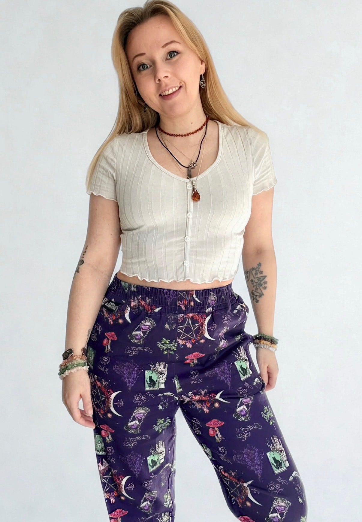 Woman wearing a white cropped top and purple patterned trousers with a mystical witchy print on a plain white wall background. Studio photo. 