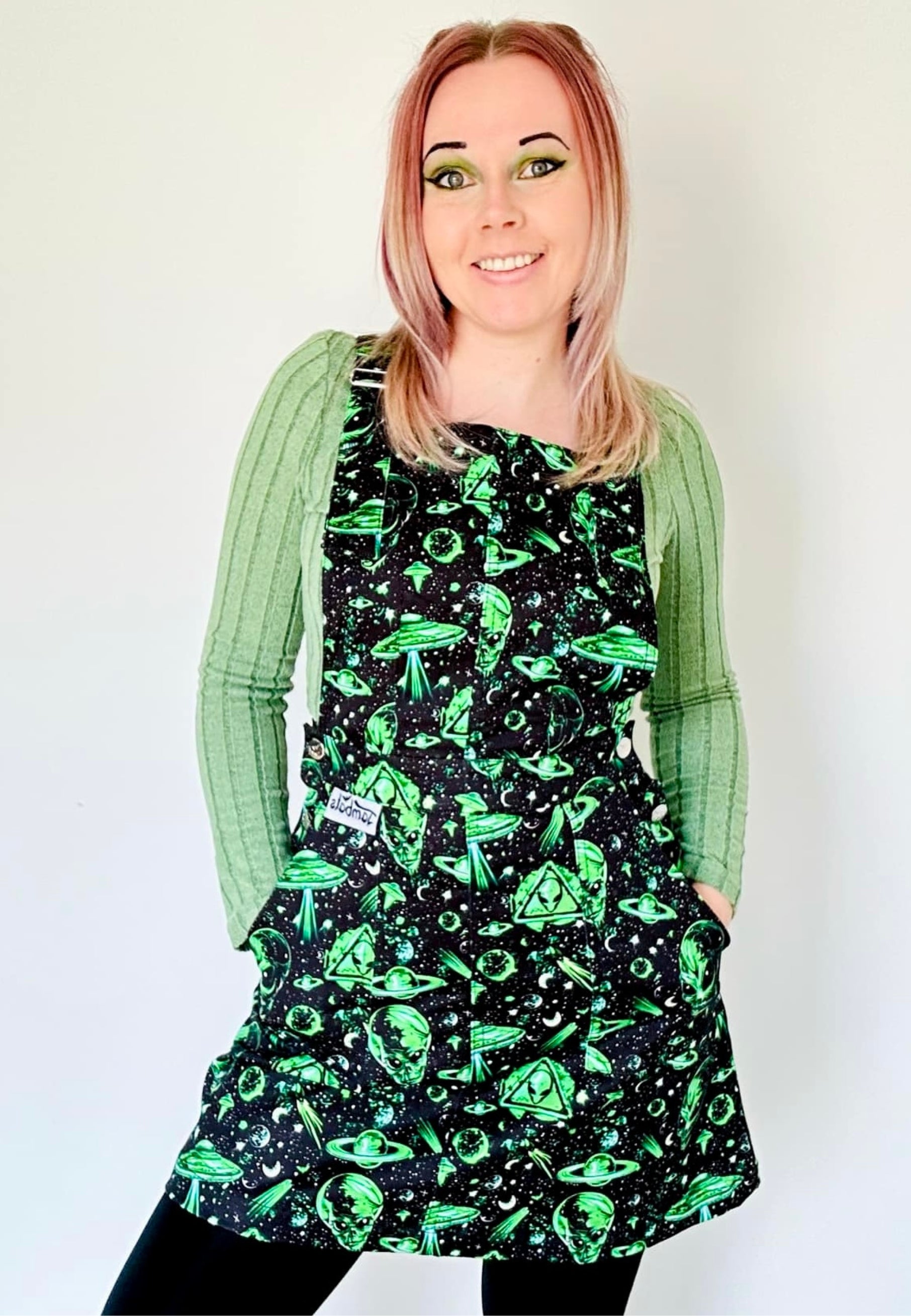 A woman wearing a black and green pinafore dress with an extraterrestrial alien space print and deep hip pockets, standing against a plain background.