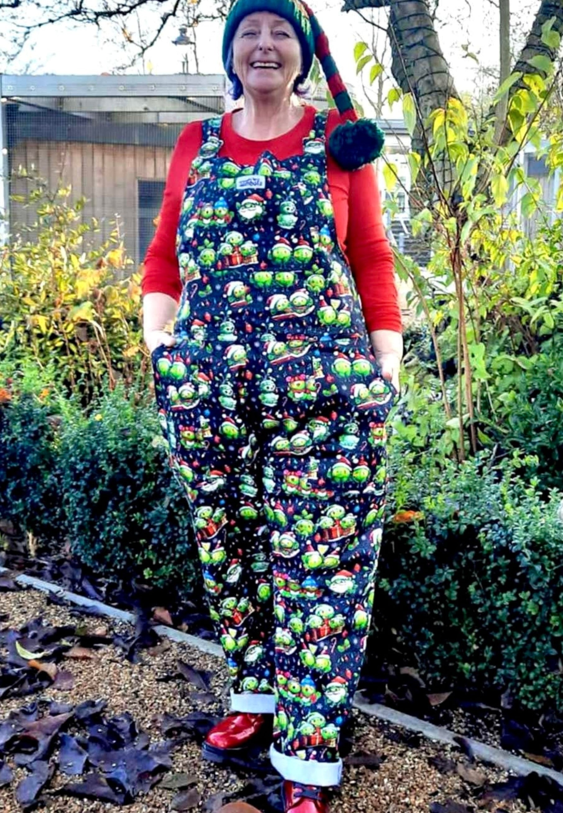 A woman wearing a festive dungarees with a pattern of Christmas sprouts. From carol singing to snowman building, Christmas partying to snow sledging… cheeky Sprouts celebrating the season. Woman is outdoors wearing a cute Santa hat and sparkly festive boots 