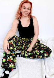 Woman wearing a black tank top and colorful patterned lounge pants sitting on a white surface. Pants have the cutest Christmas Sprouts print
