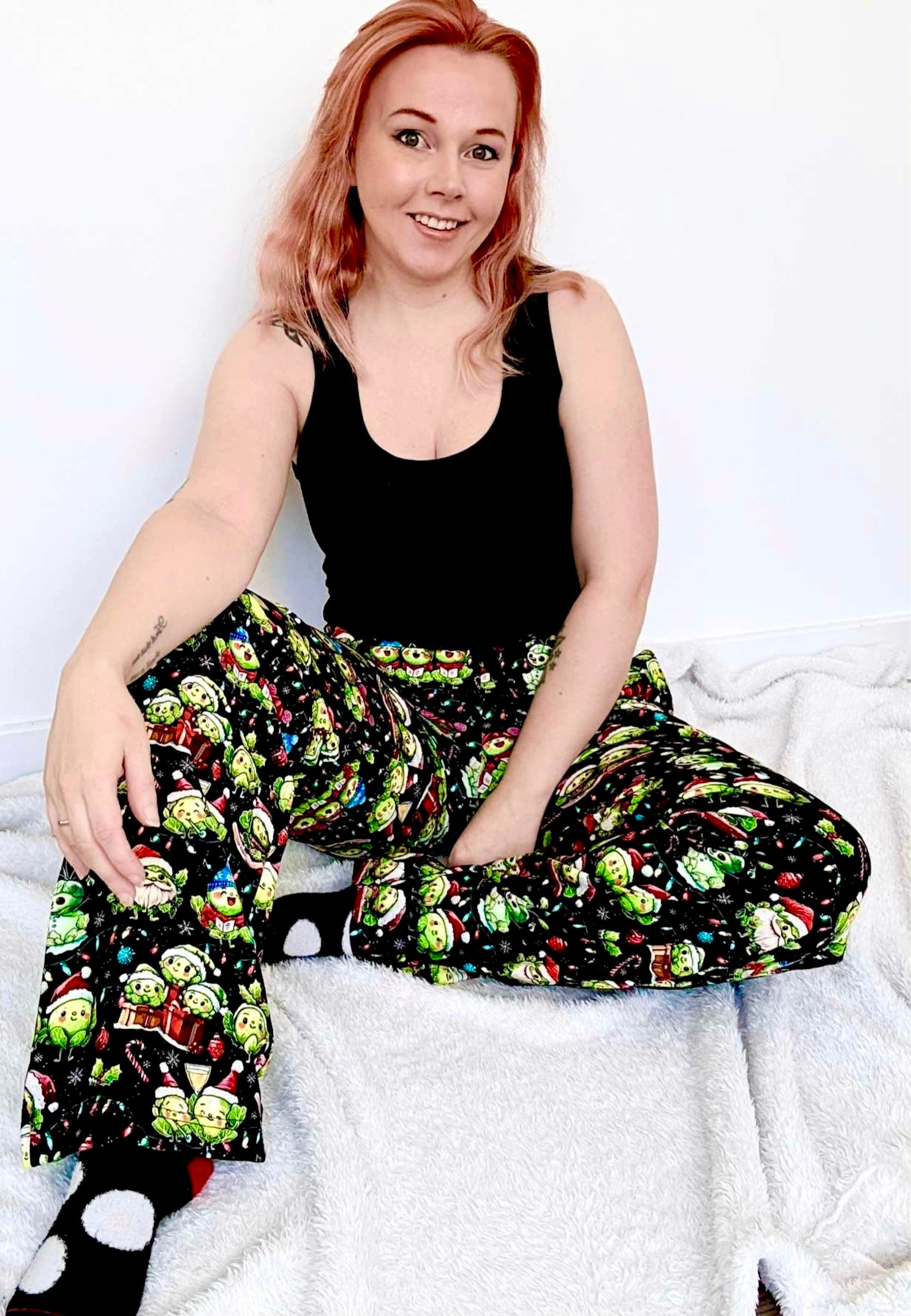 Woman wearing a black tank top and colorful patterned lounge pants sitting on a white surface. Pants have the cutest Christmas Sprouts print