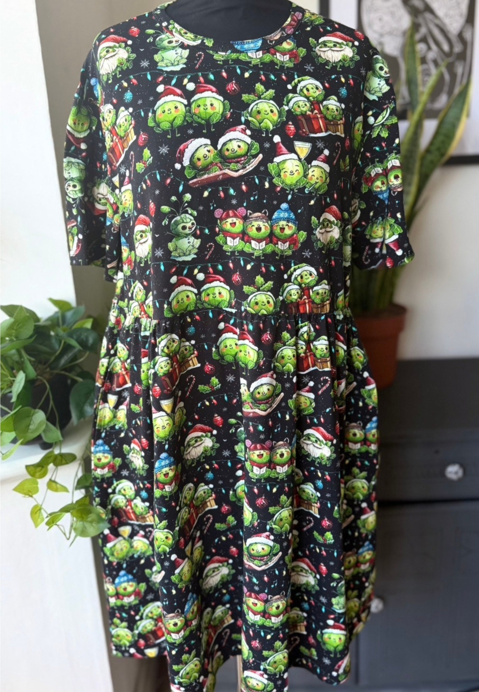 Dress with bright festive sprouts christmsd print on a black background, standing against a neutral indoor setting.