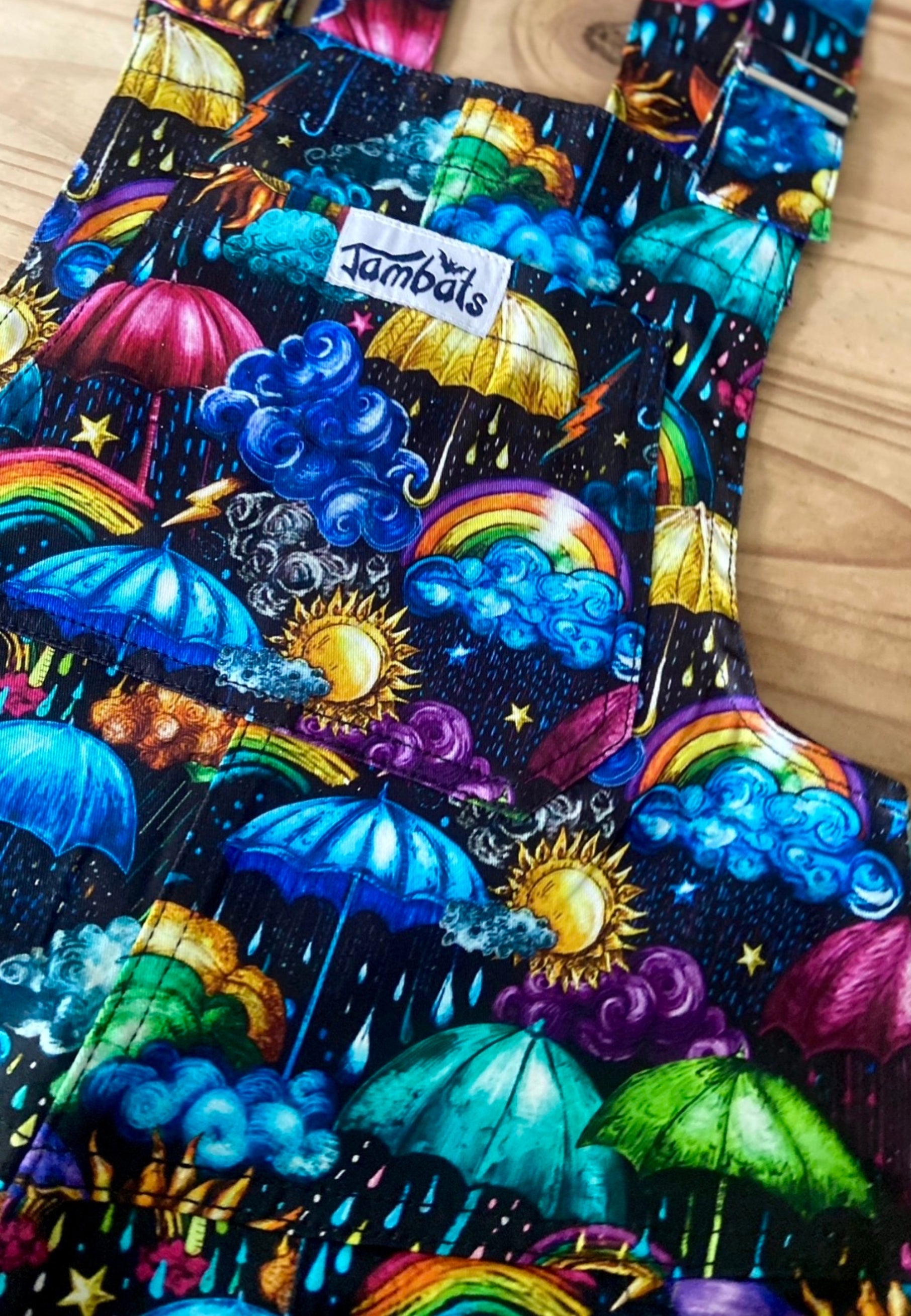Flat lay of some dungarees with a beautiful, vibrant coloured weather print. Whimsical fabric print of umbrellas, clouds, rain, sunshine, rainbows, storm clouds. 