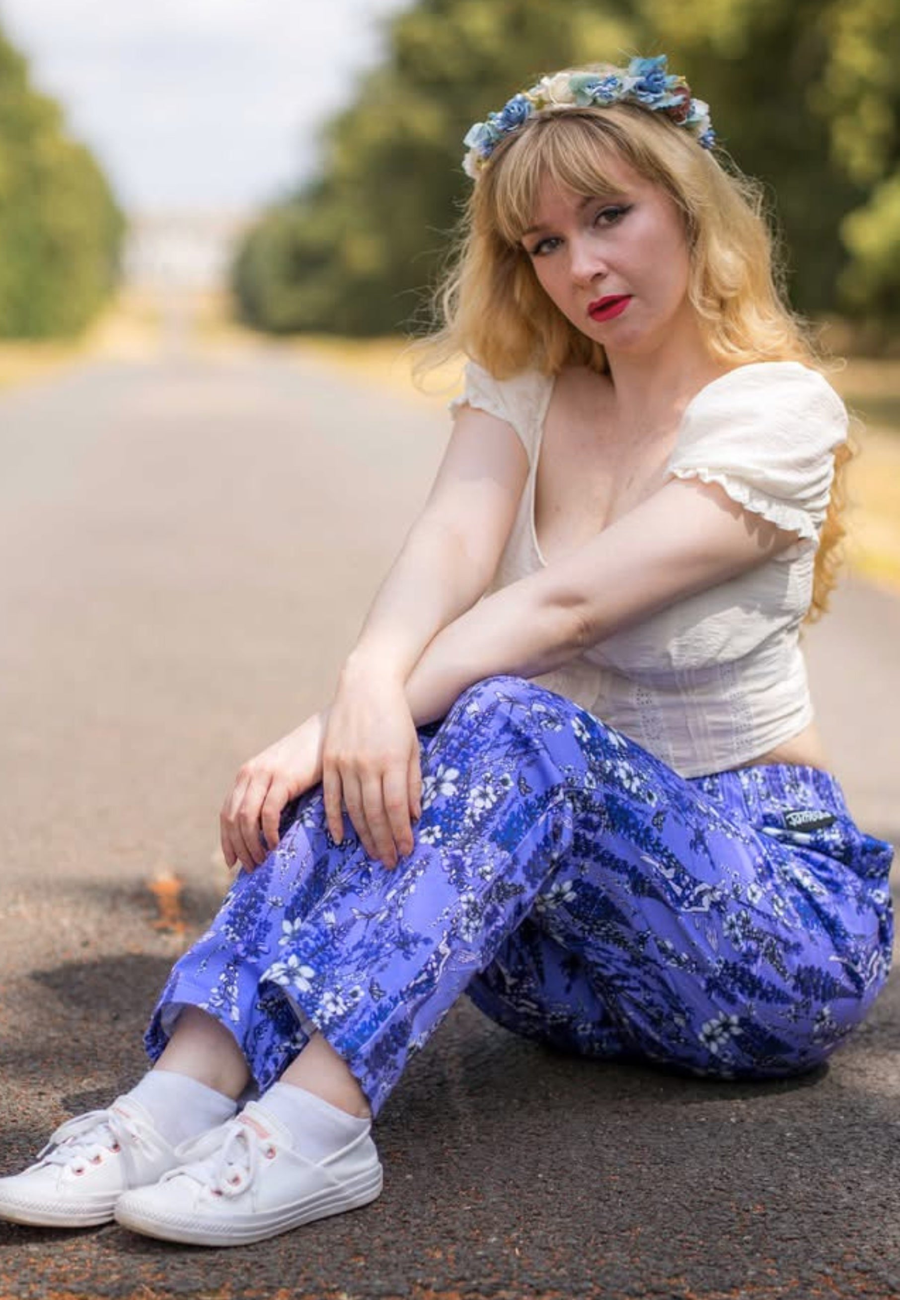 A woman sits on s road outdoors, wearing a white ruffled short-sleeve top, purple floral trousers and white pumps. 