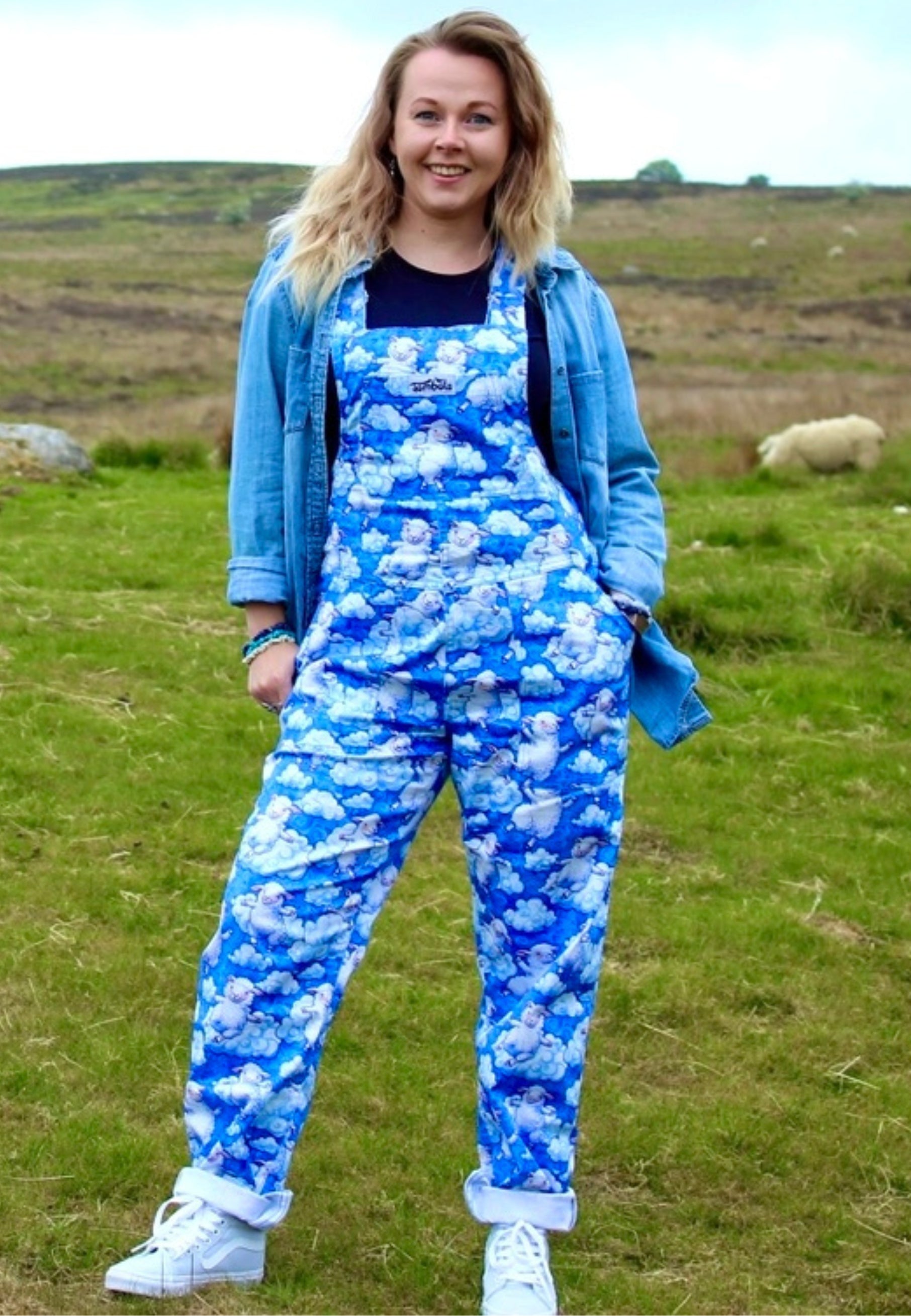 Model wearing a blue fluffy cloud and fluffy sheep patterned dungarees standing in a grassy field with sheep in the background. 