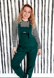 A woman wearing forest green dungarees and a black and white striped top against a quirky striped wall background