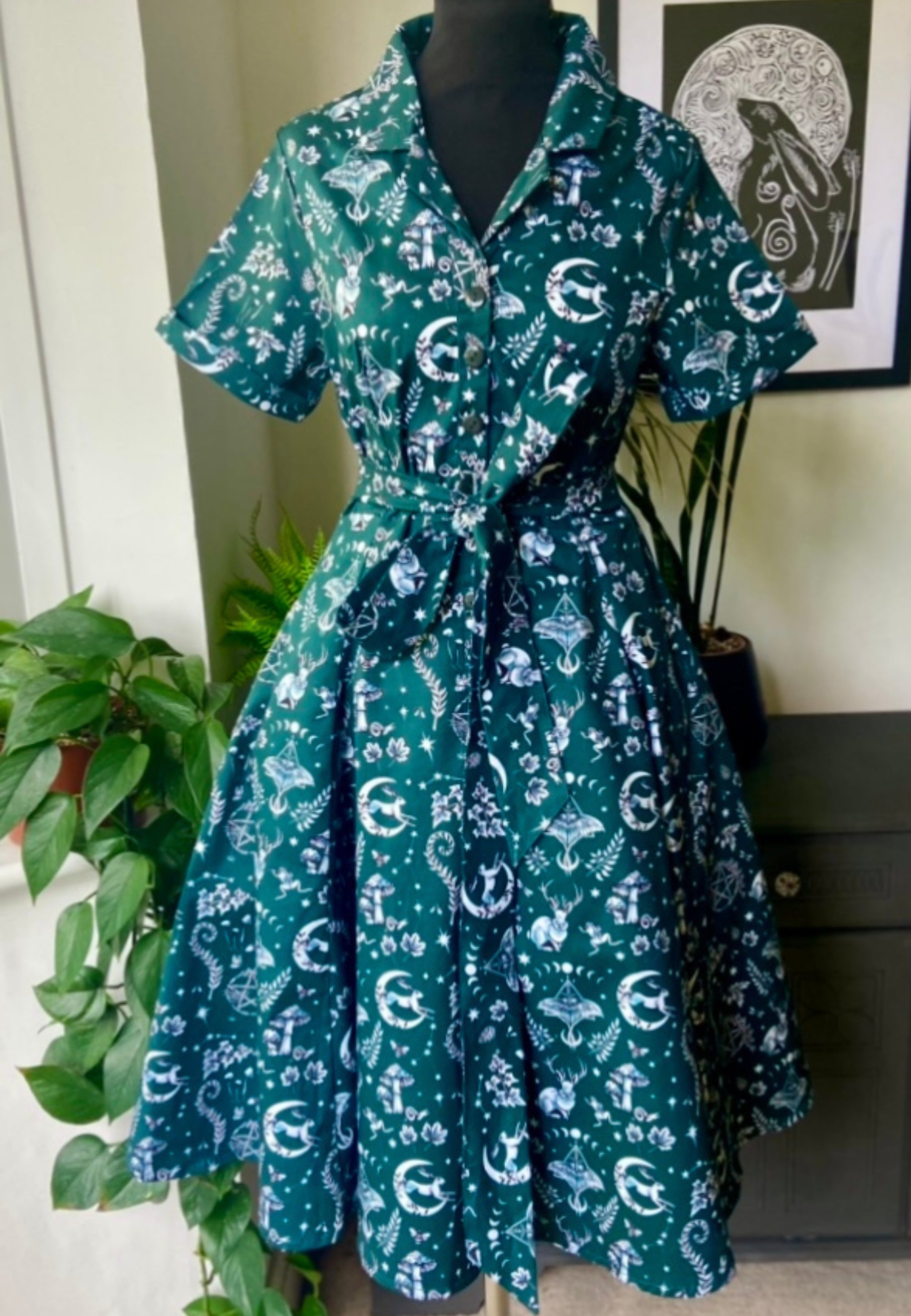 A deep forest green retro shirt dress with short sleeves, button-up shirt bodice, and a full skirt. It features a unique illustrated magick and nature-themed print, including elements like moons, rabbits, and constellations.