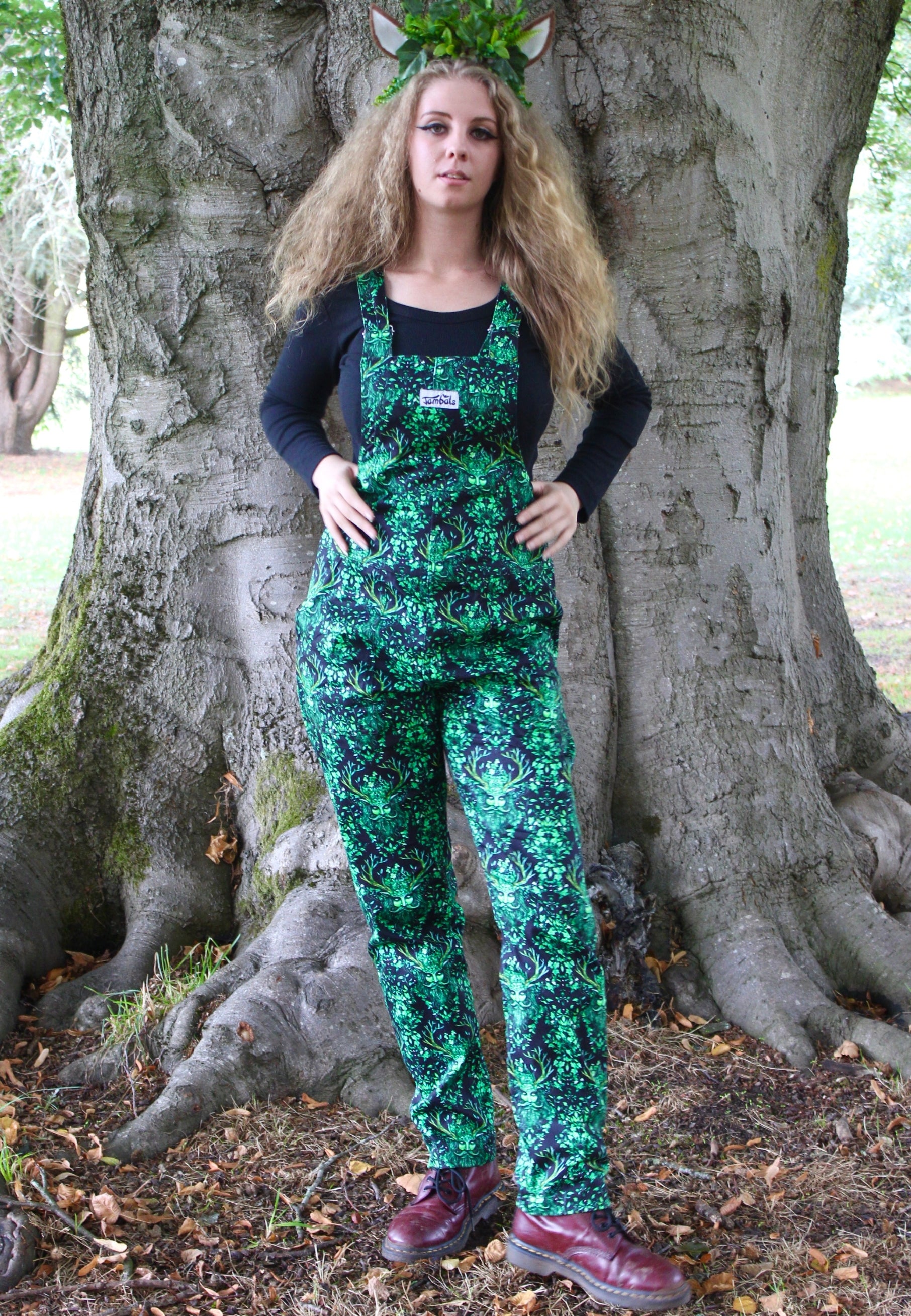 Woman standing in a forest setting wearing black dungarees with a beautiful green man print on the dungarees. Witchy pagan style.